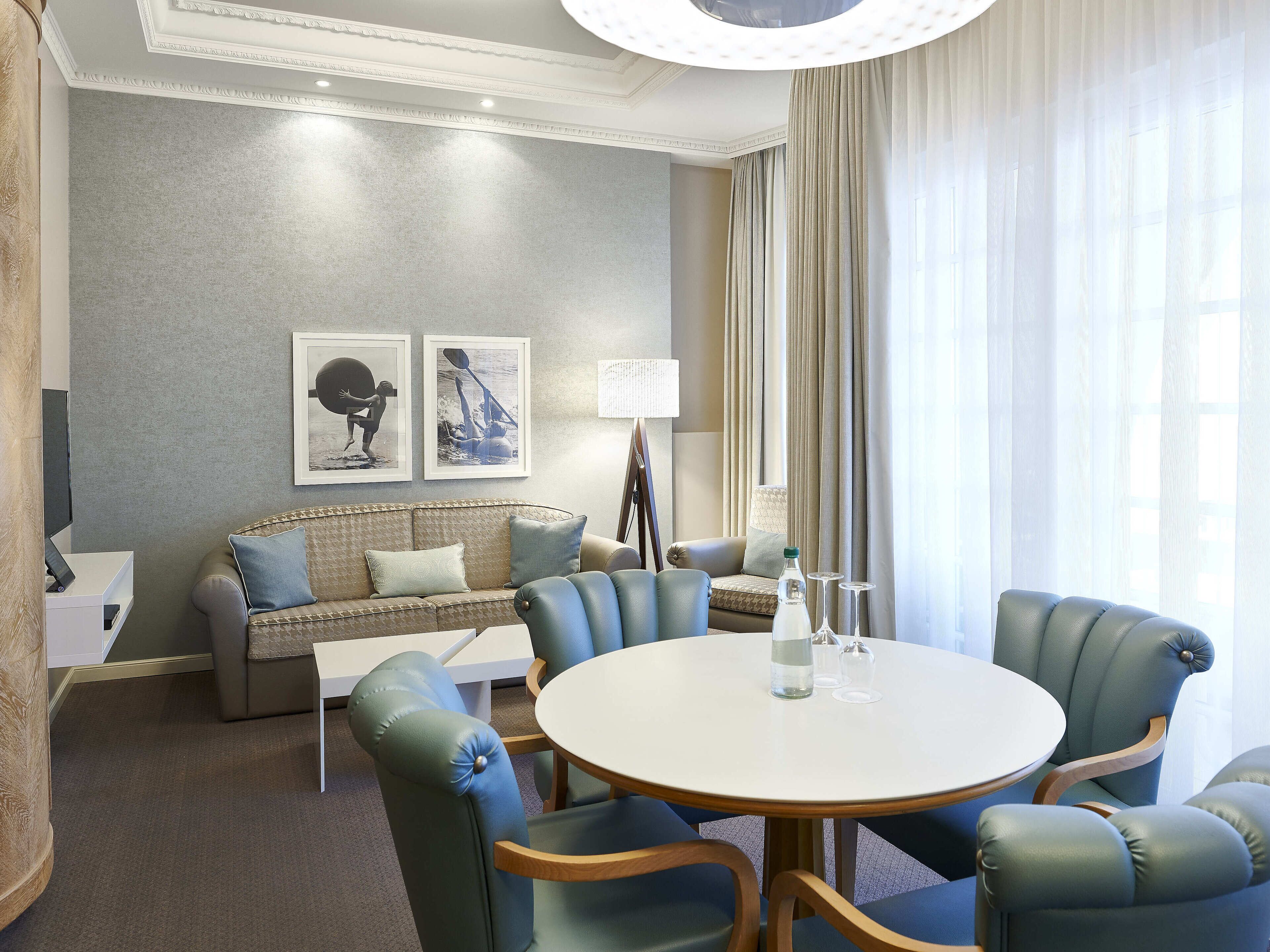 A modern hotel room with a grey textured wall, a beige sofa with blue cushions, a round white table with chairs, and a large window with sheer curtains.