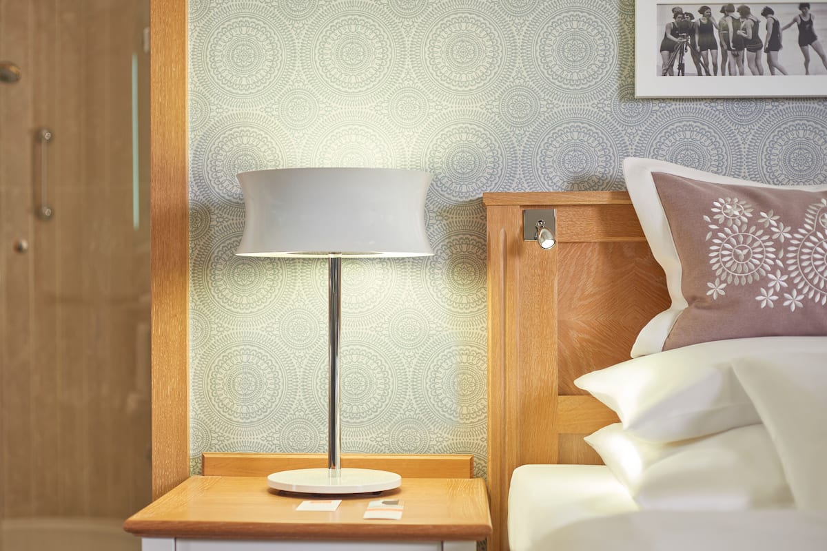 A neatly made bed with a white comforter and a decorative pillow, a wooden nightstand with a lamp, and patterned wallpaper.