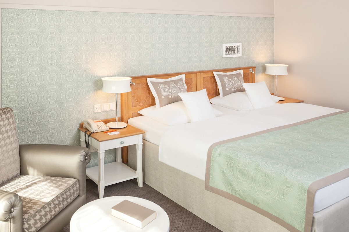 A hotel room with a bed, bedside tables, a chair, and a small table with a book.