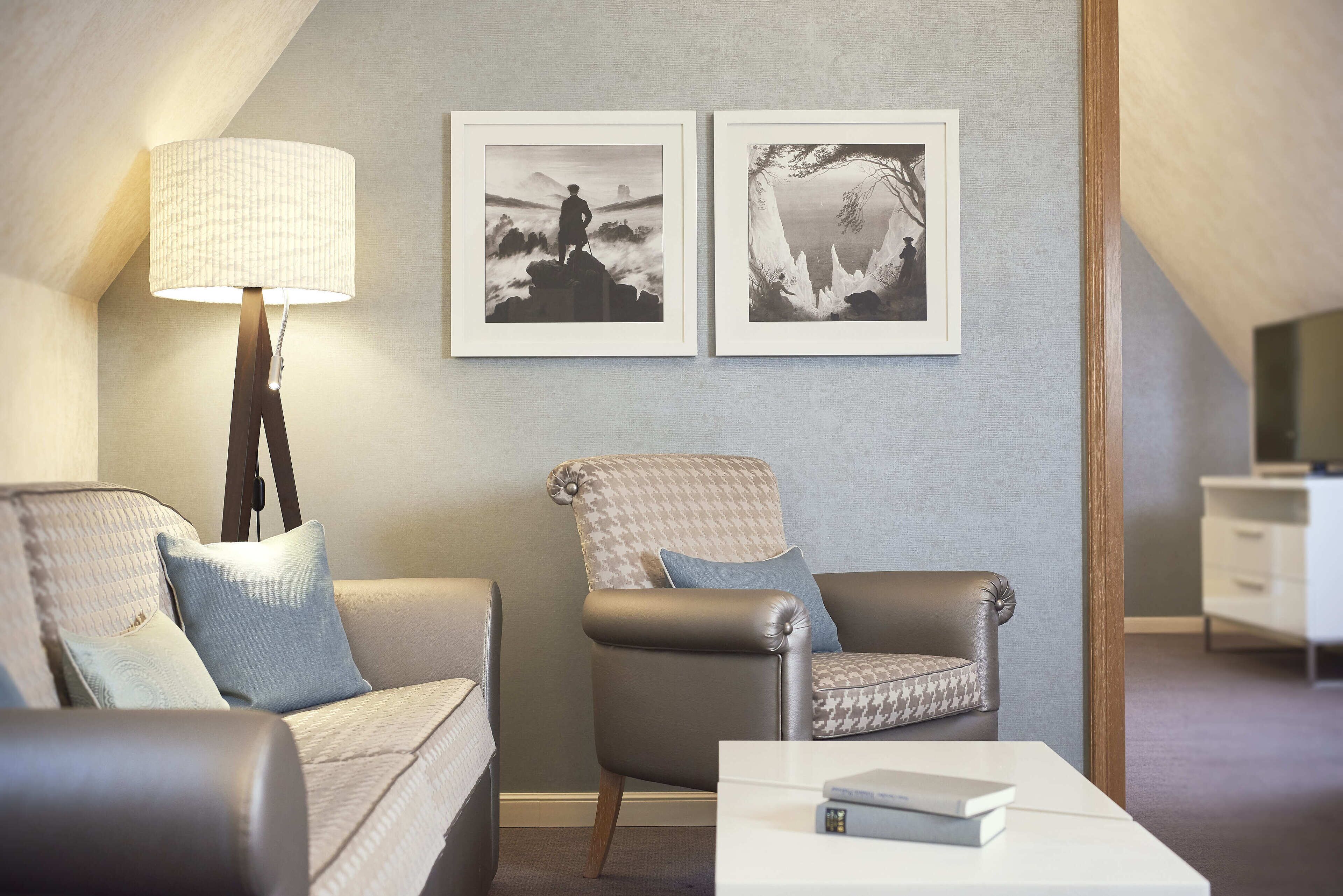 Ein modernes Wohnzimmer mit einer Couch, einem Sessel und einer Lampe. An der Wand hängen zwei gerahmte Bilder.