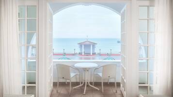 A room with a view of the sea, a small building, and two chairs at a table.