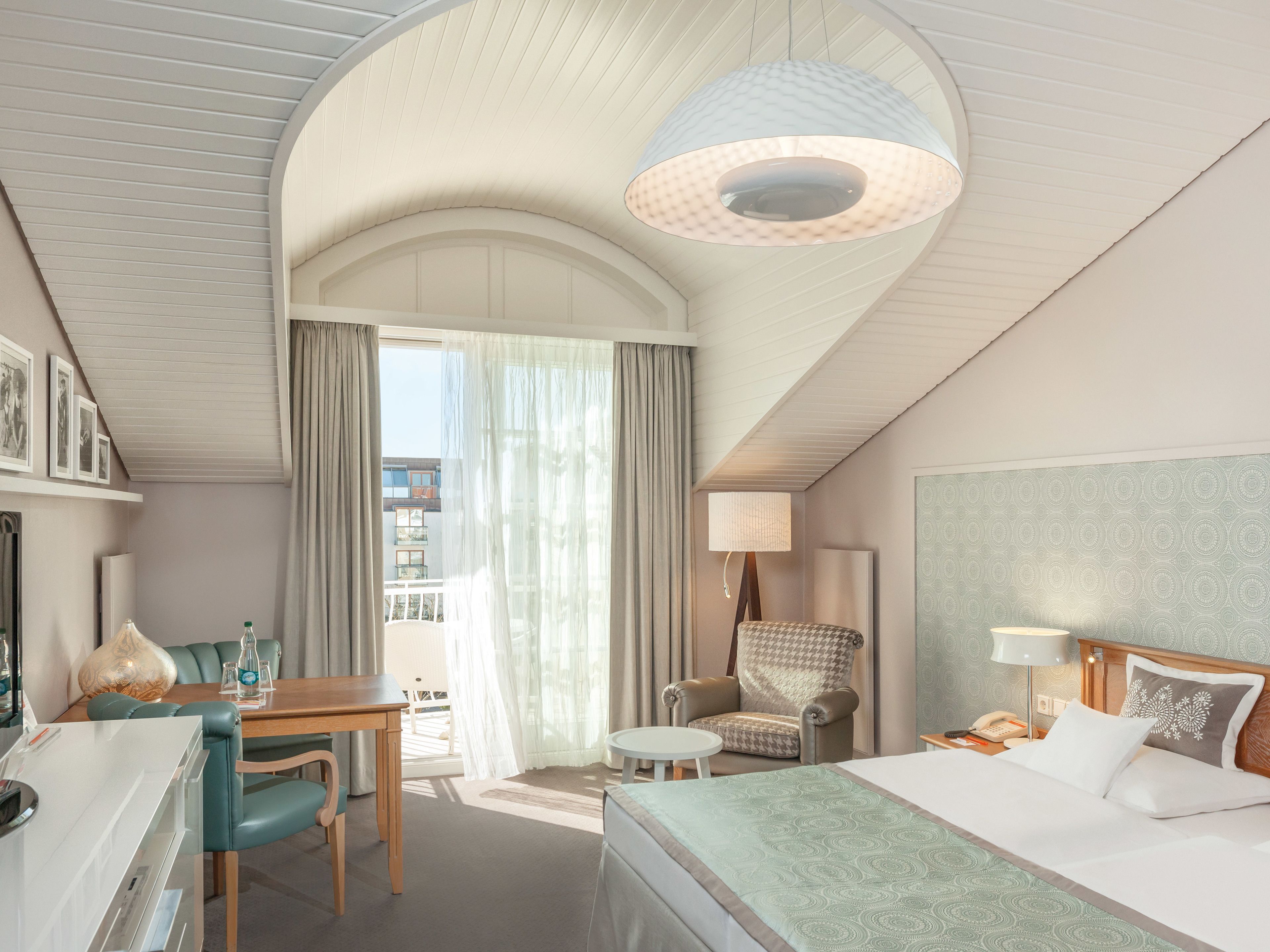 A hotel room with a bed, a desk, a chair, a table, and a balcony with curtains.