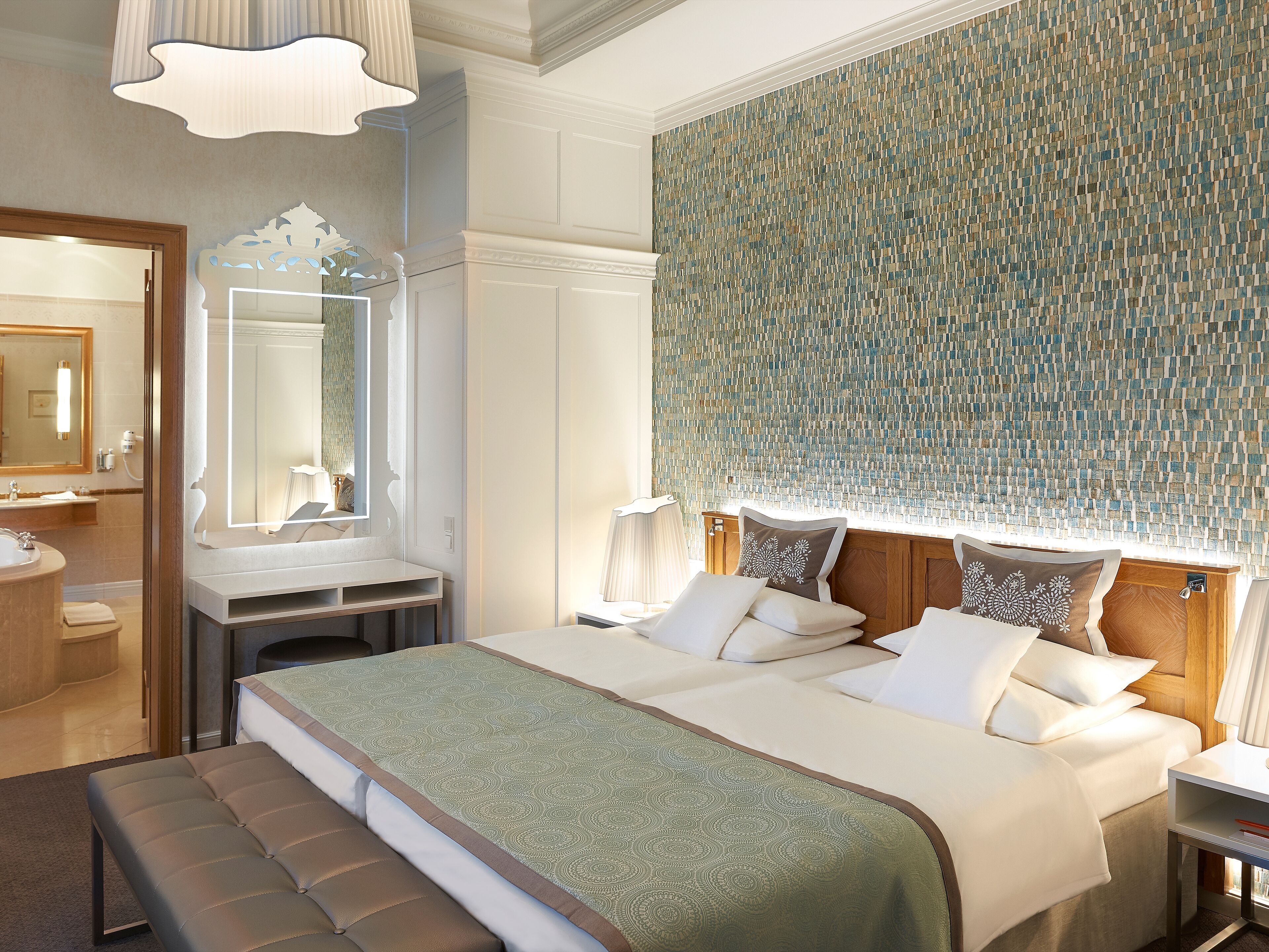 A hotel room with a large bed, a bedside table, a mirror, a bathroom with a sink and toilet, and a patterned wall.