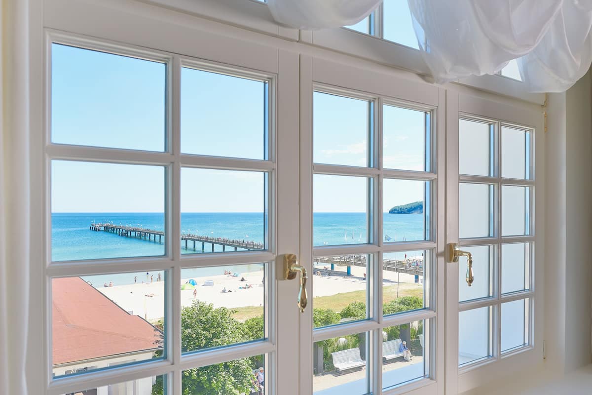 A view through a window with white frames, revealing a beach, a pier, and a clear blue sky.