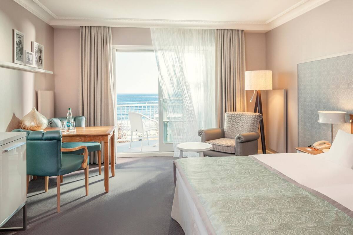 A hotel room with a bed, a desk with chairs, a small table, a lamp, and a view of the ocean.