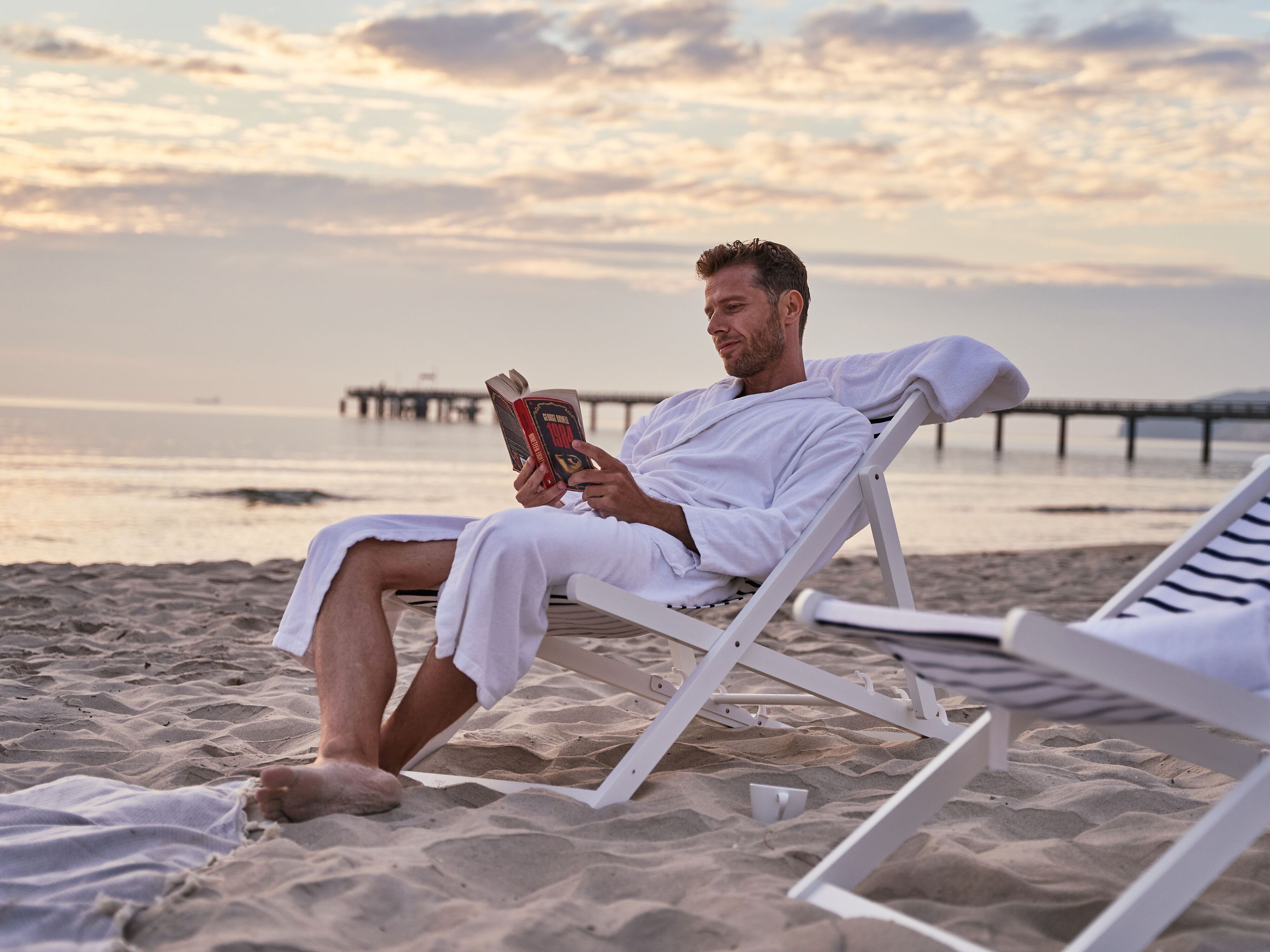 Uma pessoa lendo um livro em uma cadeira de praia, com vista para o oceano e um píer ao fundo.