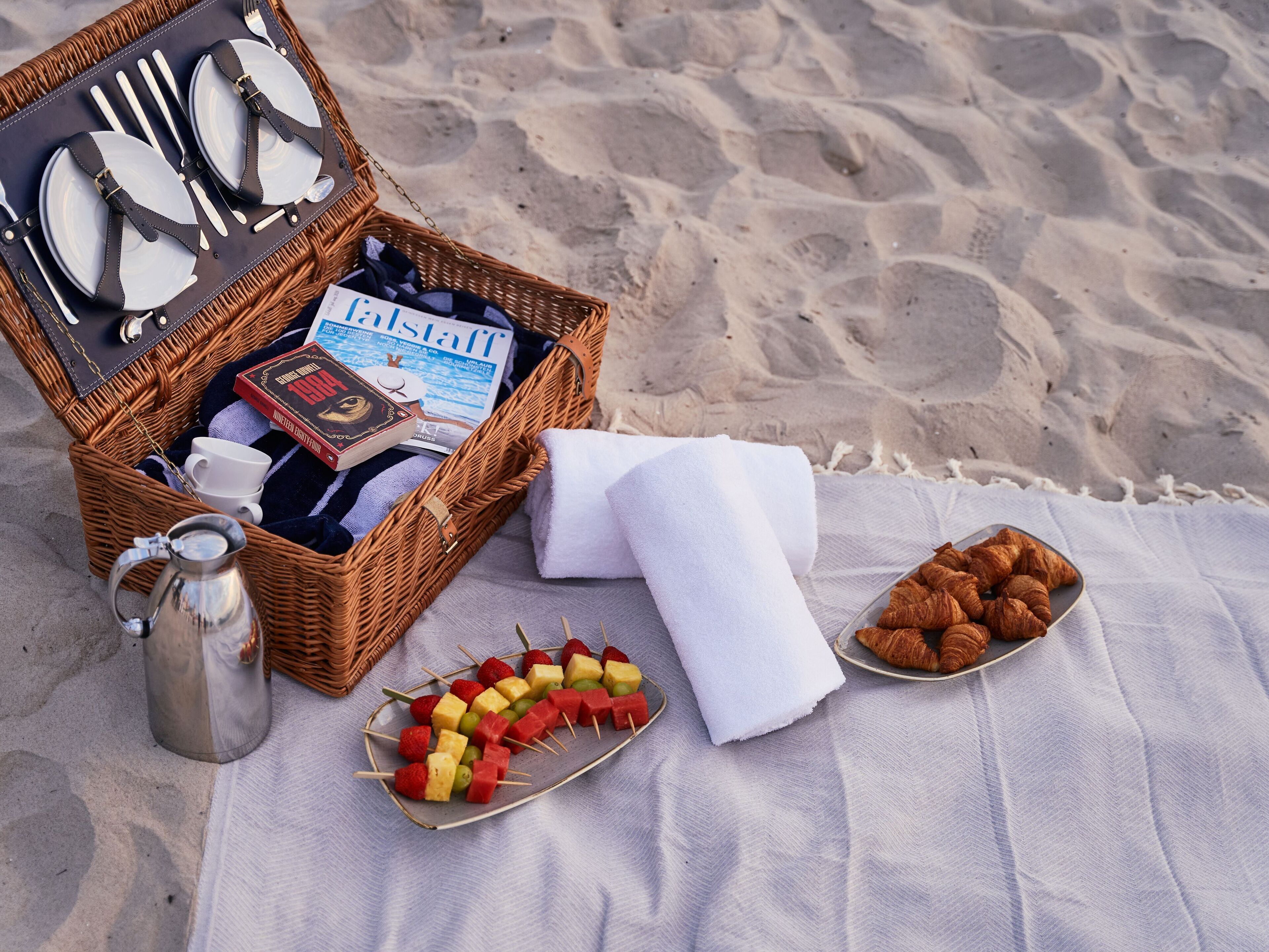 Een picknick op het strand met een rieten mand die een boek, een mok en een thermos bevat. Op een deken liggen spiesjes met fruit en croissants.