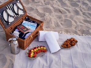 A beach picnic setup with a wicker basket containing a book, a mug, and a thermos. There are skewers of fruit and croissants on a blanket. - A-ROSA  Kurhaus Binz (Binz)