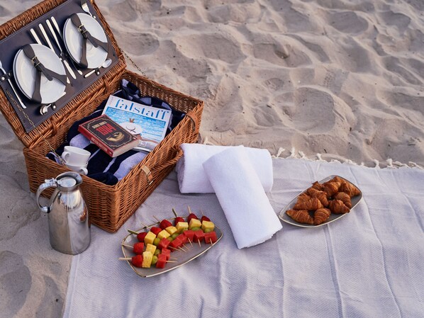 A beach picnic setup with a wicker basket containing a book, a mug, and a thermos. There are skewers of fruit and croissants on a blanket.