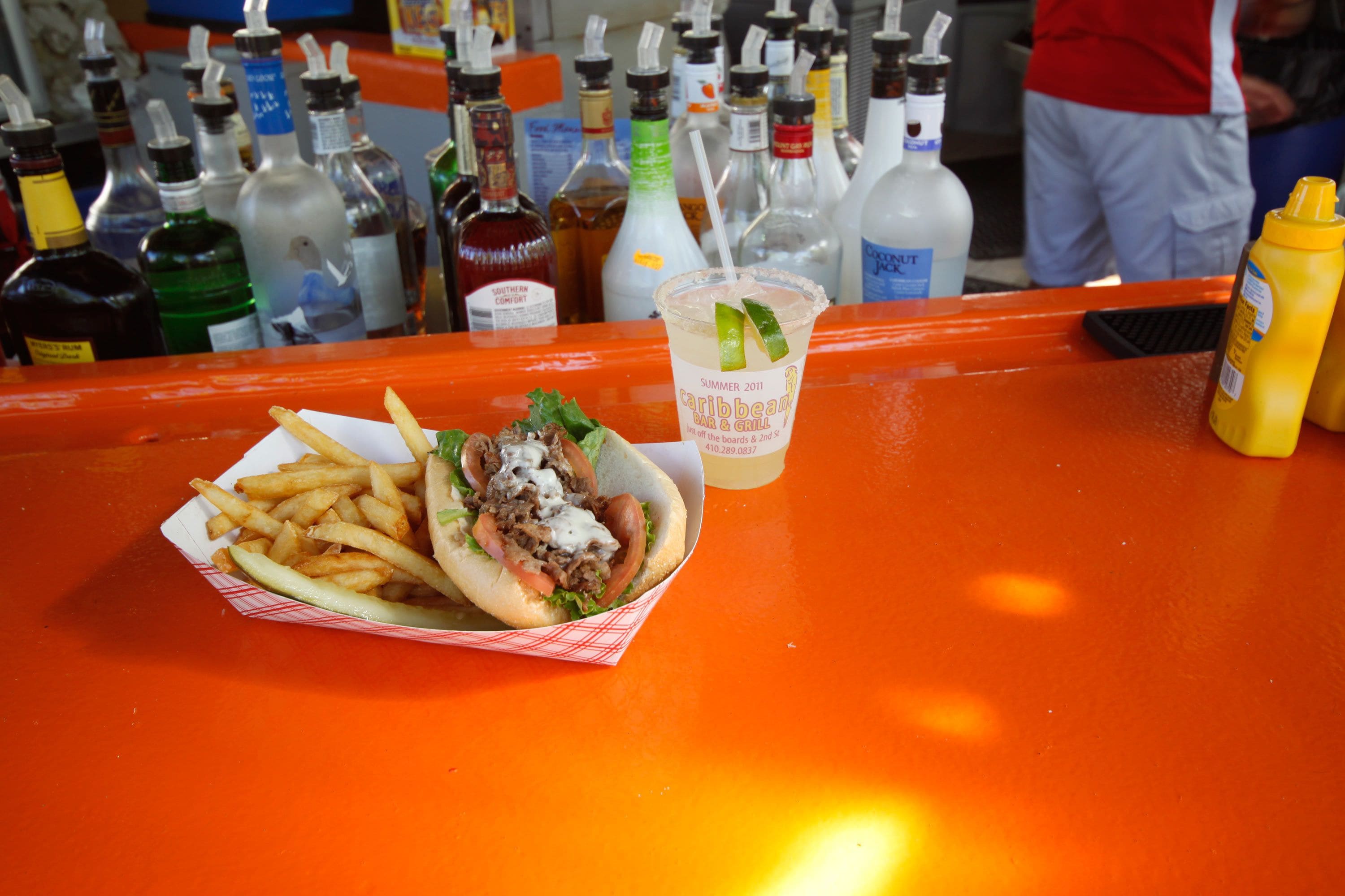 Comida que incluye un sándwich y papas fritas sobre una mesa naranja, con una bebida y varias botellas al fondo.