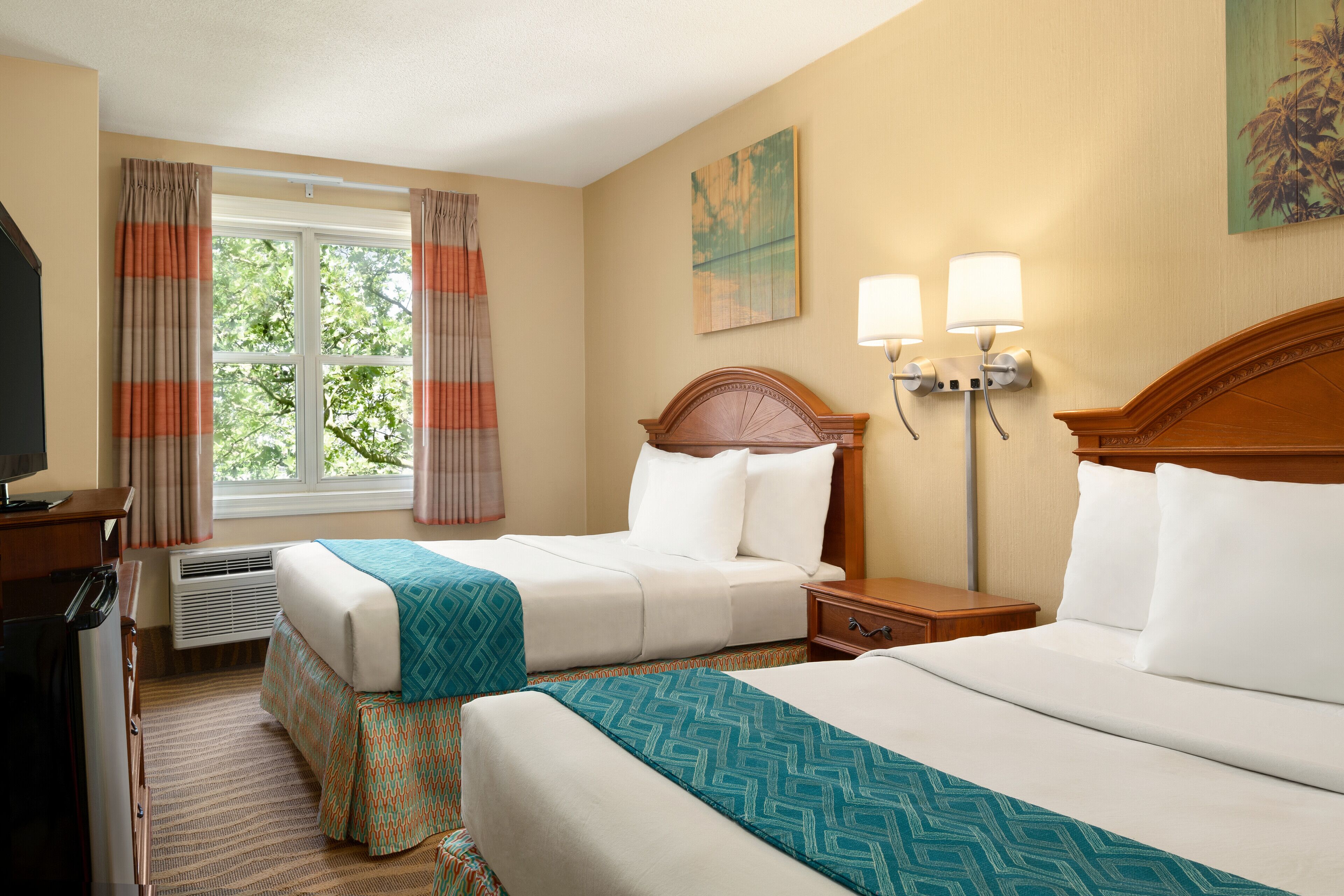 A hotel room with two beds, a desk with a computer, a window with curtains, and wall-mounted lamps.