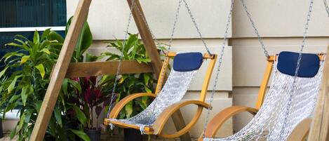 Una silla colgante de madera con asiento y respaldo tipo hamaca, suspendida por cadenas, frente a una pared de edificio.