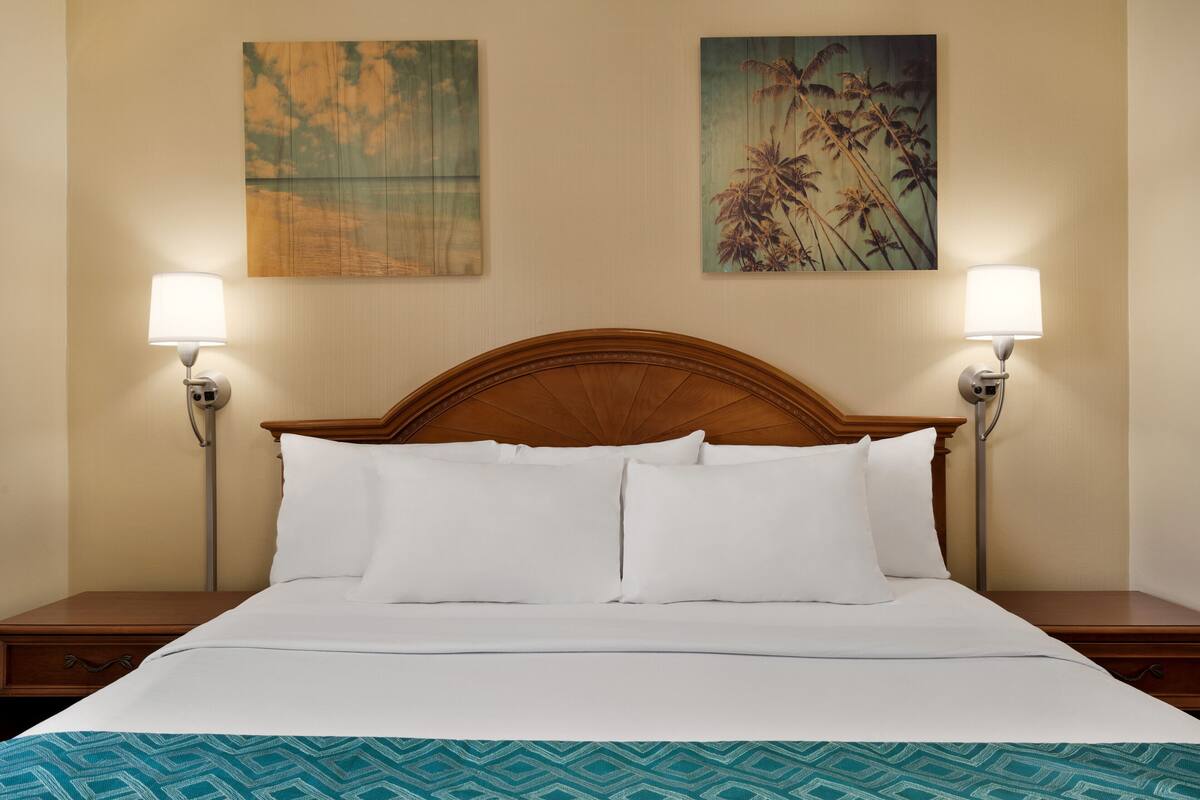 A neatly made bed with white linens, flanked by two bedside tables with lamps, in a room with a wooden headboard and two wall-mounted paintings.