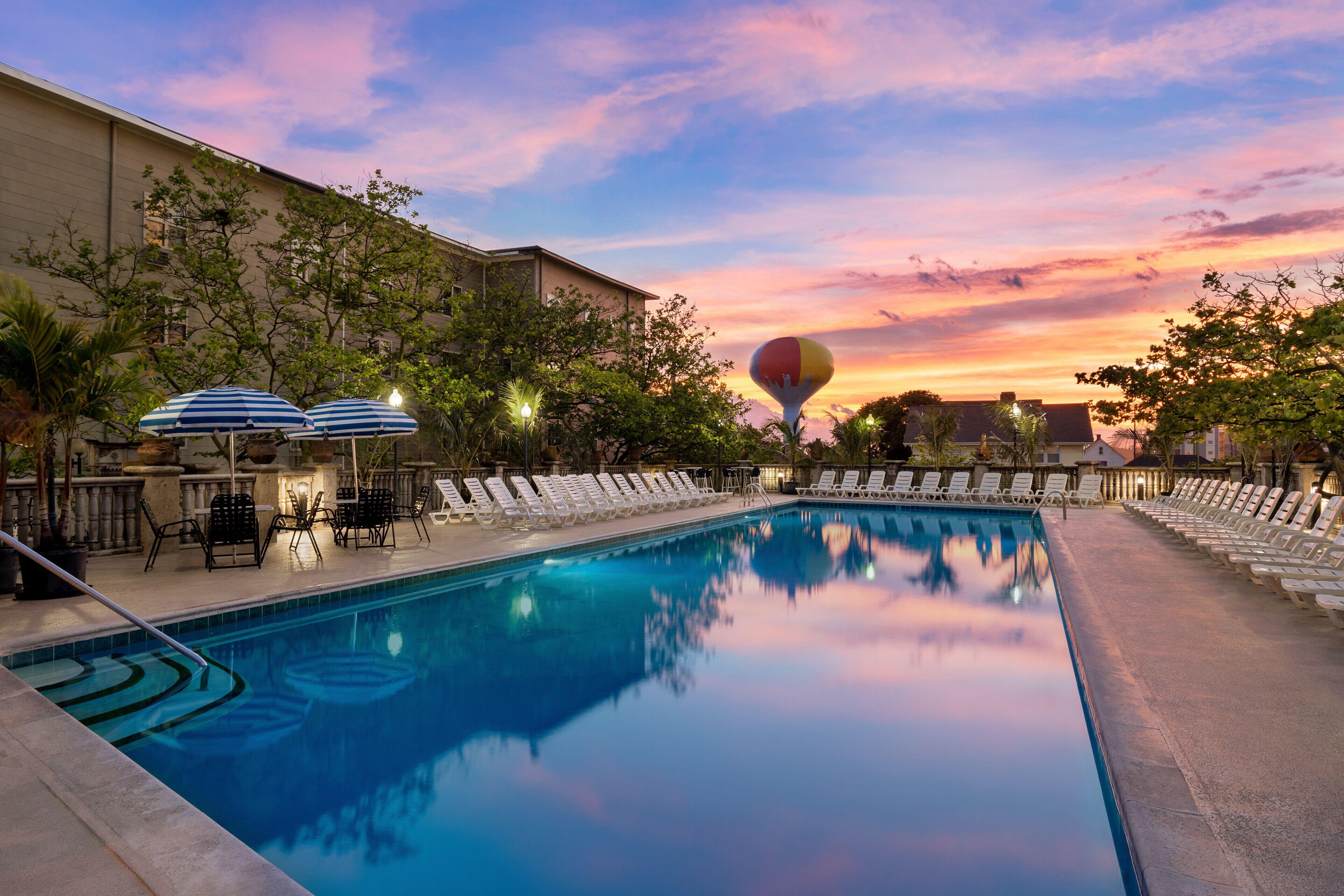 A swimming pool with lounge chairs and umbrellas, surrounded by trees and a building.