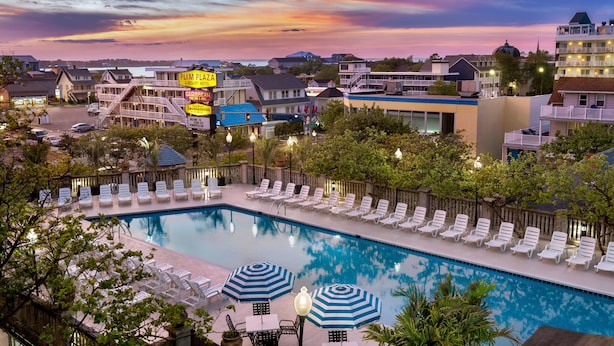 A swimming pool with lounge chairs and umbrellas, surrounded by buildings and a sunset sky. - Plim Plaza Hotel (Ocean City)