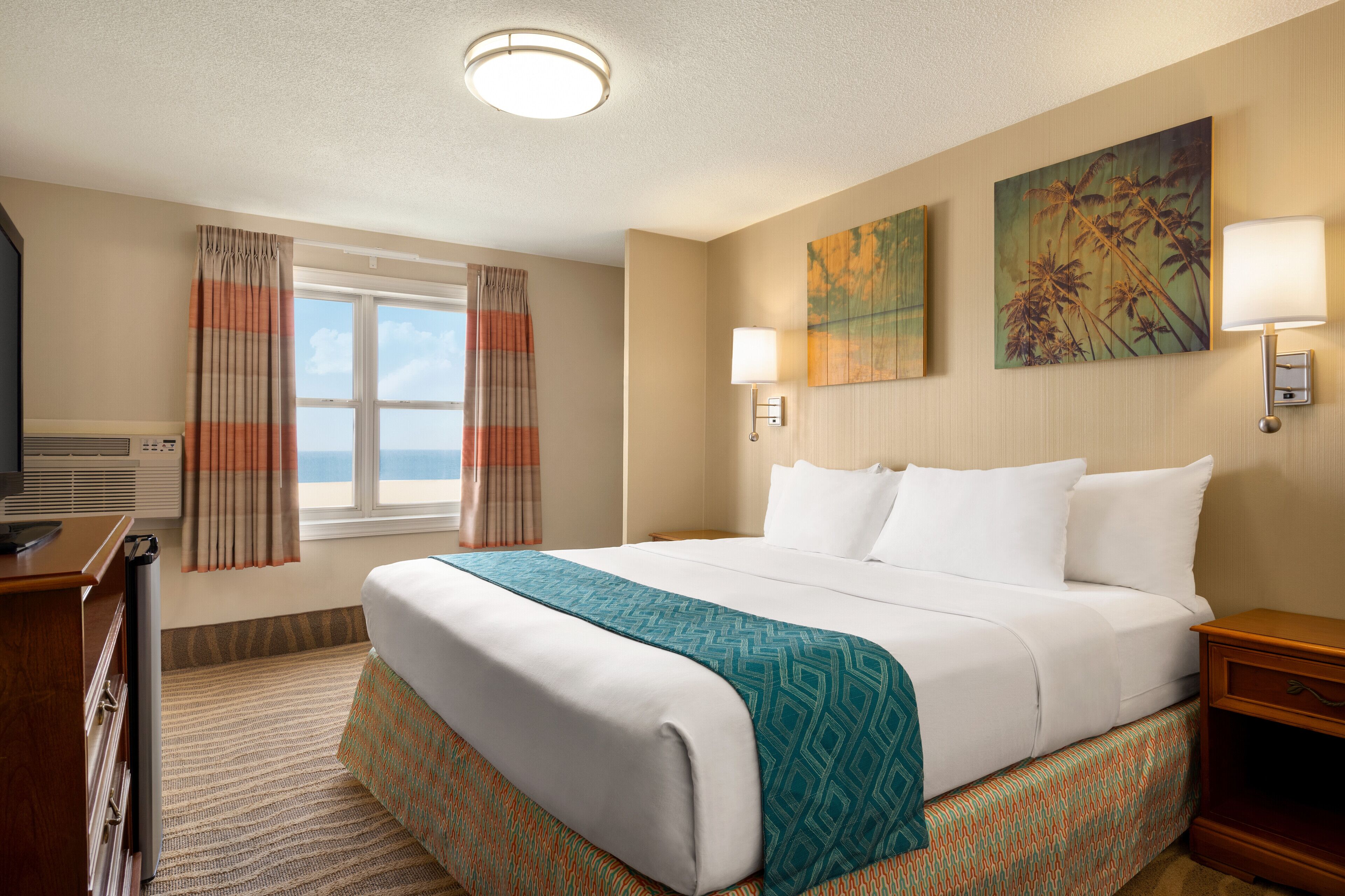 A hotel room with a large bed, a desk, and a window with curtains. There are two wall-mounted lamps and a painting on the wall.