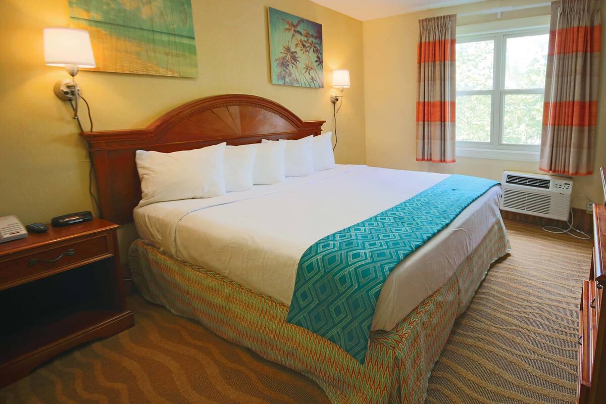 A hotel room with a large bed, two bedside lamps, a nightstand, a desk, an air conditioner, and a window with curtains.