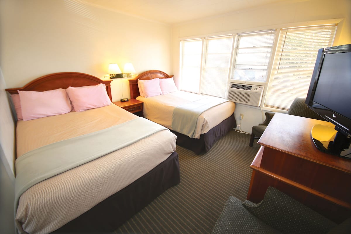 A hotel room with two beds, a desk with a television, and an air conditioning unit.
