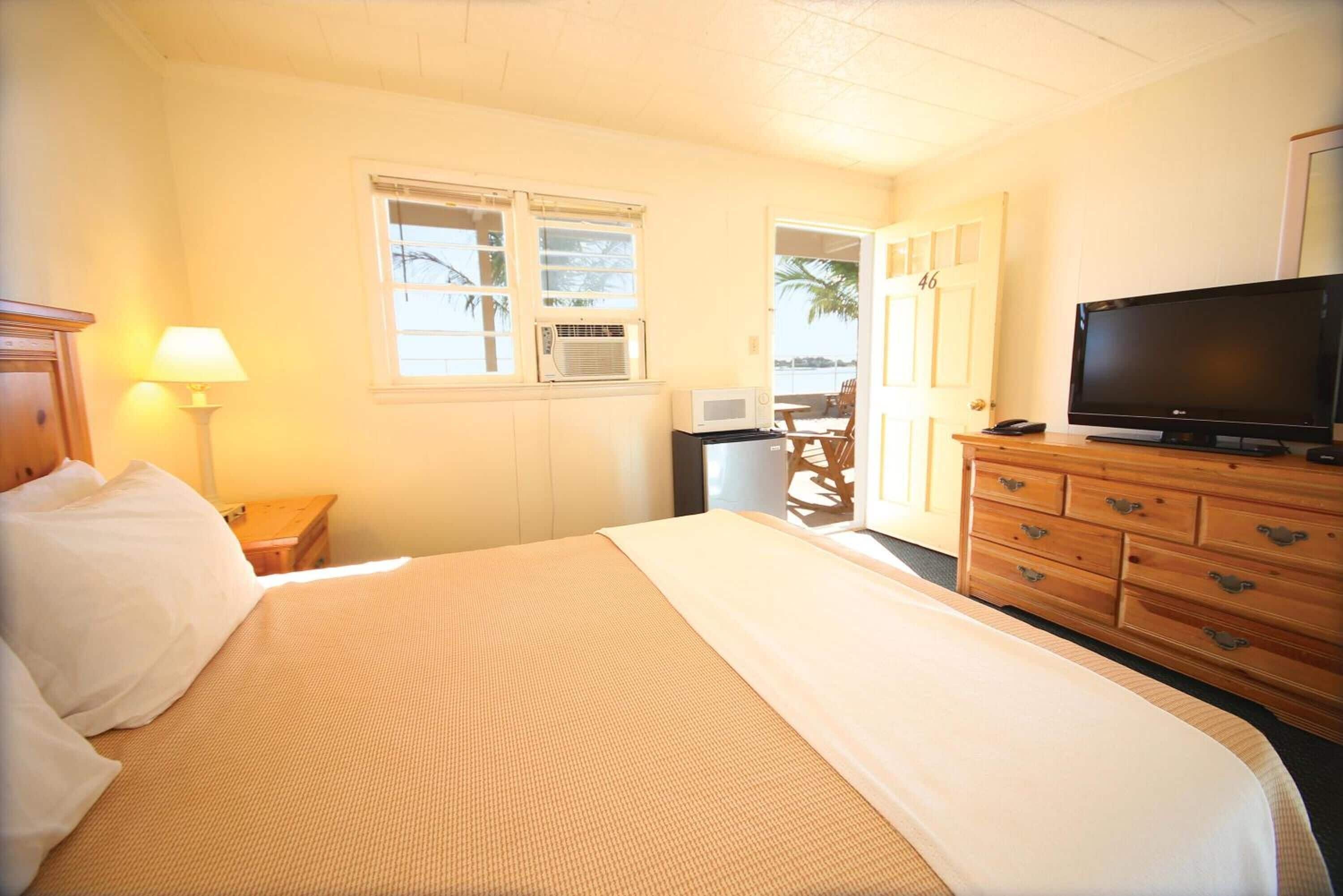 A hotel room with a bed, a television, a dresser, a lamp, and a window with an air conditioning unit.