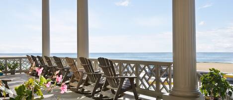 Een overdekt terras met houten stoelen en een potplant, met uitzicht op een strand.