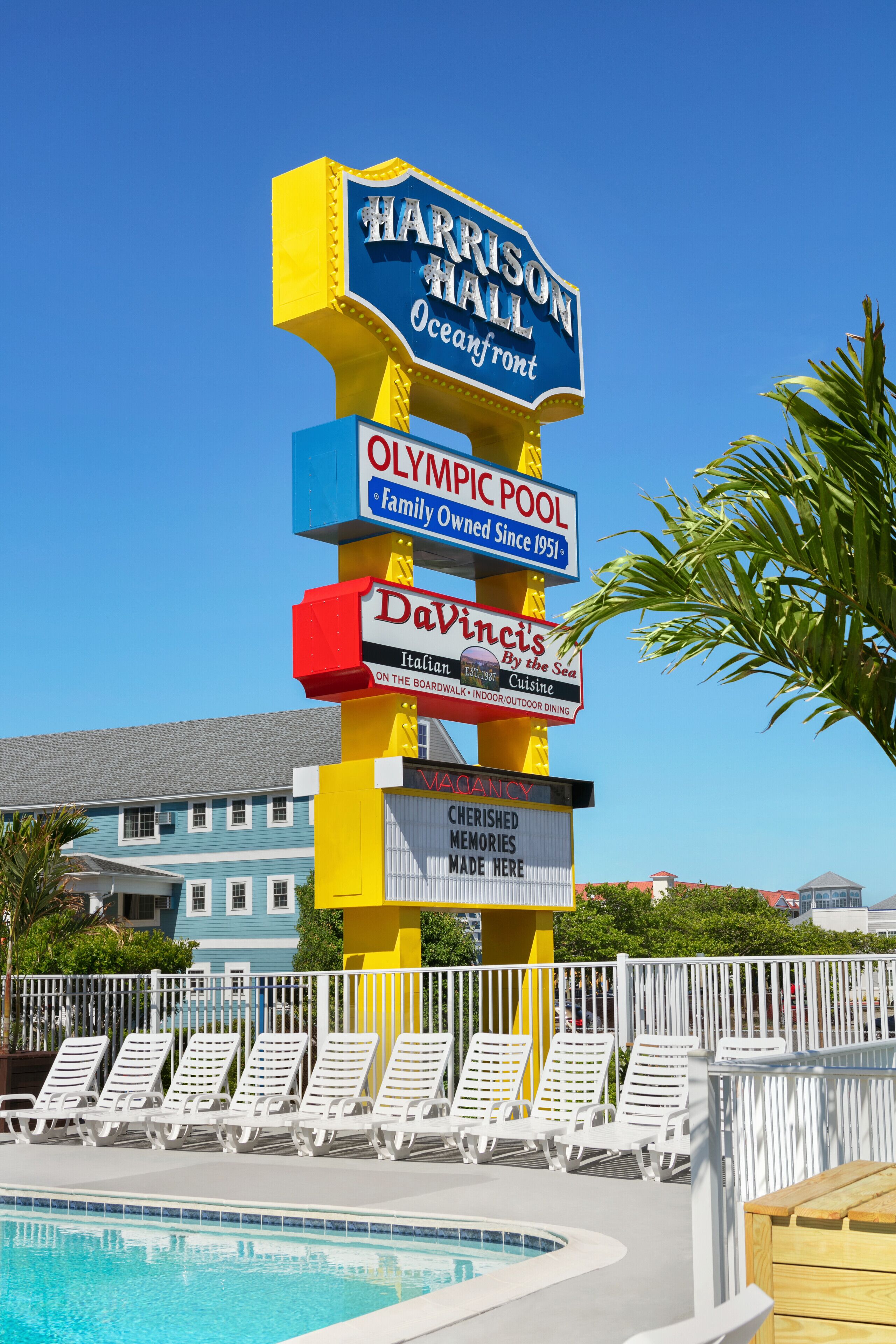 A pool area with lounge chairs, a sign for Harrison Hall Oceanfront, Olympic Pool, Davinci's Italian cuisine, and a statement about cherished memories made here.