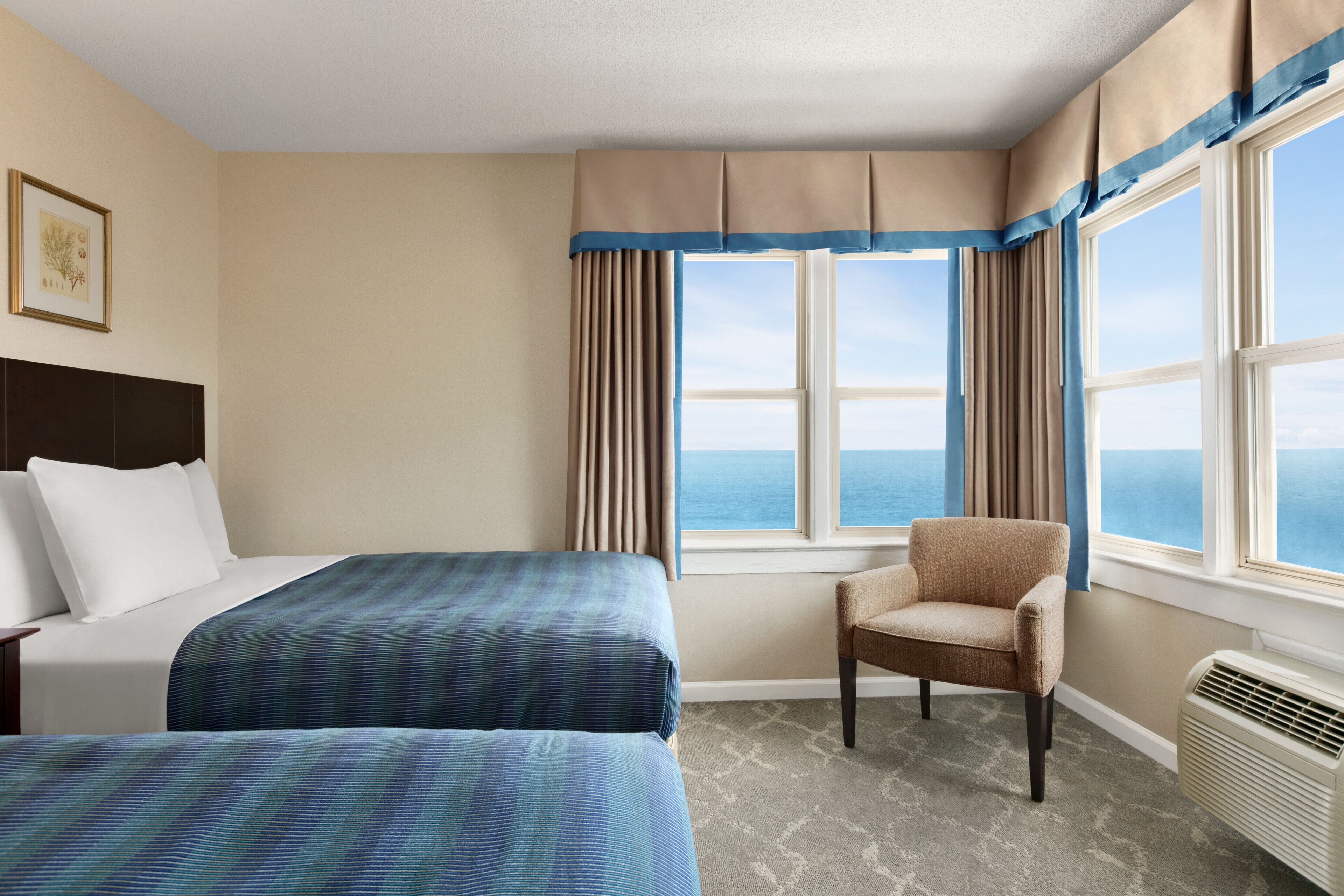 Ein Hotelzimmer mit zwei Betten, einem Sessel, einem großen Fenster mit Meerblick und einem Deckenventilator.