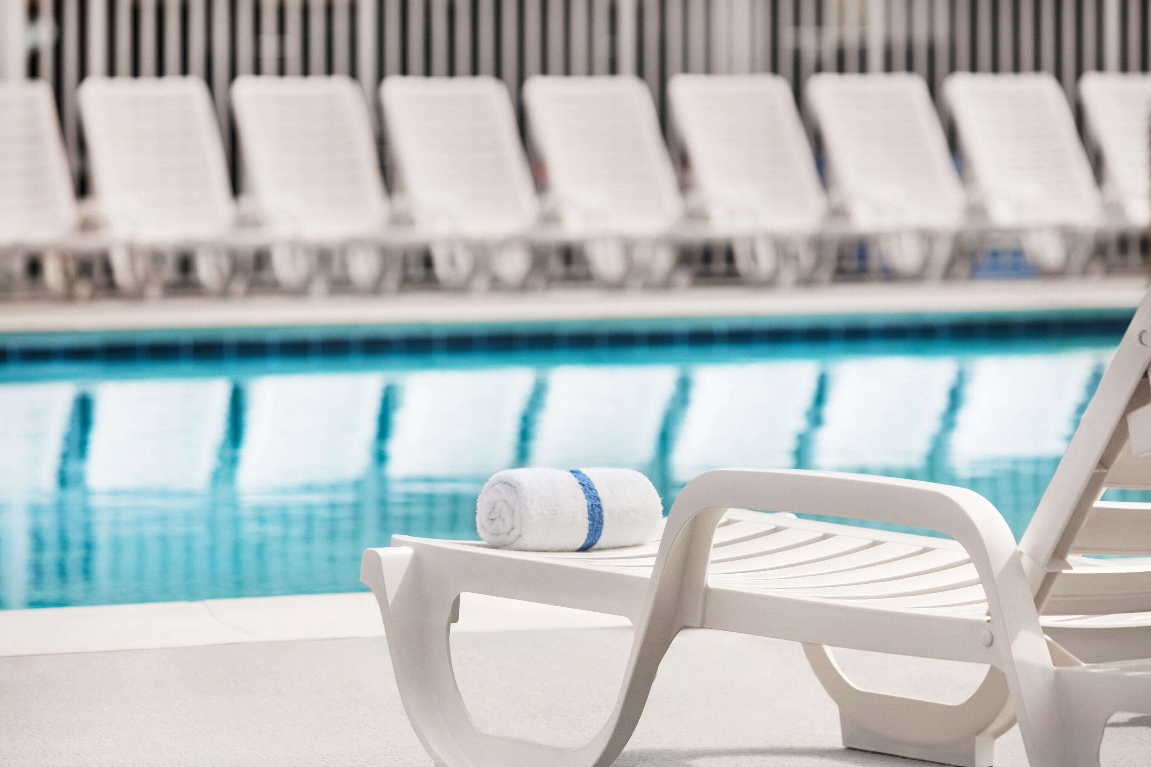 A poolside area with white lounge chairs and a rolled towel on one of them.