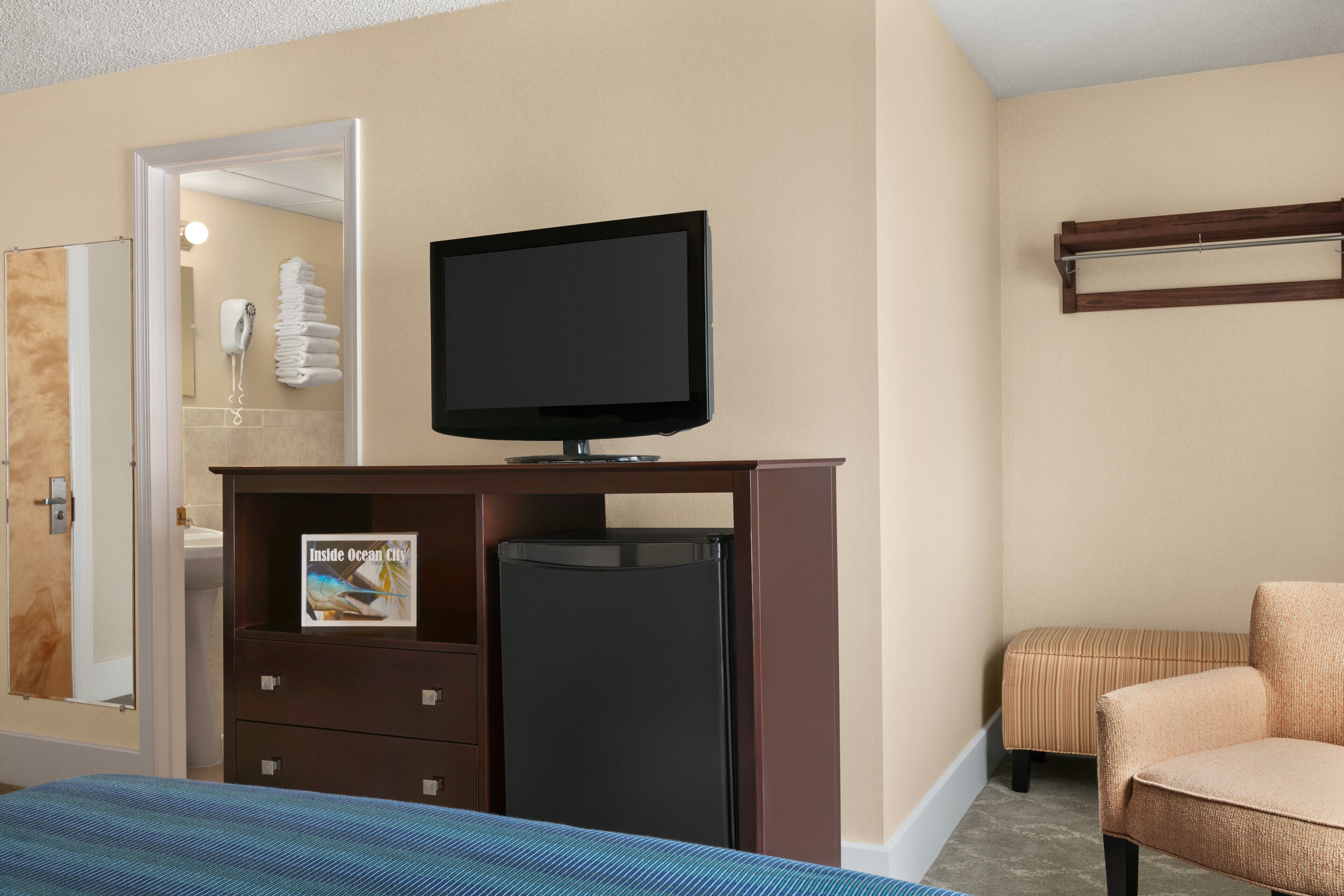Habitación de hotel con cama, televisor sobre un mueble de madera, refrigerador, baño con toallero y una zona de estar con sillón y reposapiés.