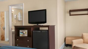 Habitación de hotel con cama, televisor sobre un mueble de madera, refrigerador, baño con toallero y una zona de estar con sillón y reposapiés.