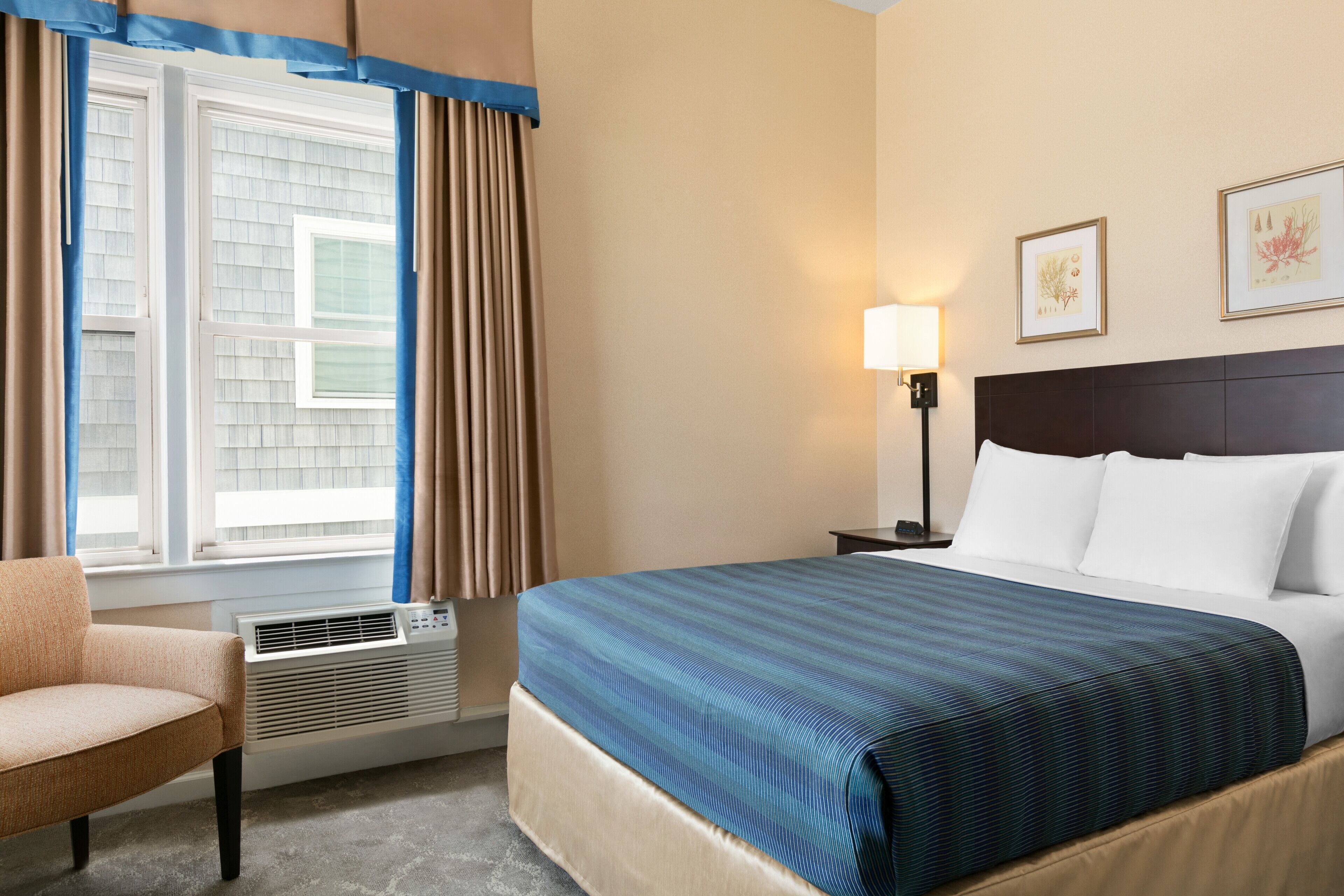 A hotel room with a bed, a chair, a window with curtains, a wall-mounted lamp, and framed pictures.