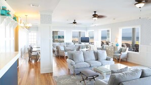 A spacious living room with a large sofa, a coffee table, and a view of the beach. There are ceiling fans and a TV in the room. - Harrison Hall Hotel (Ocean City)