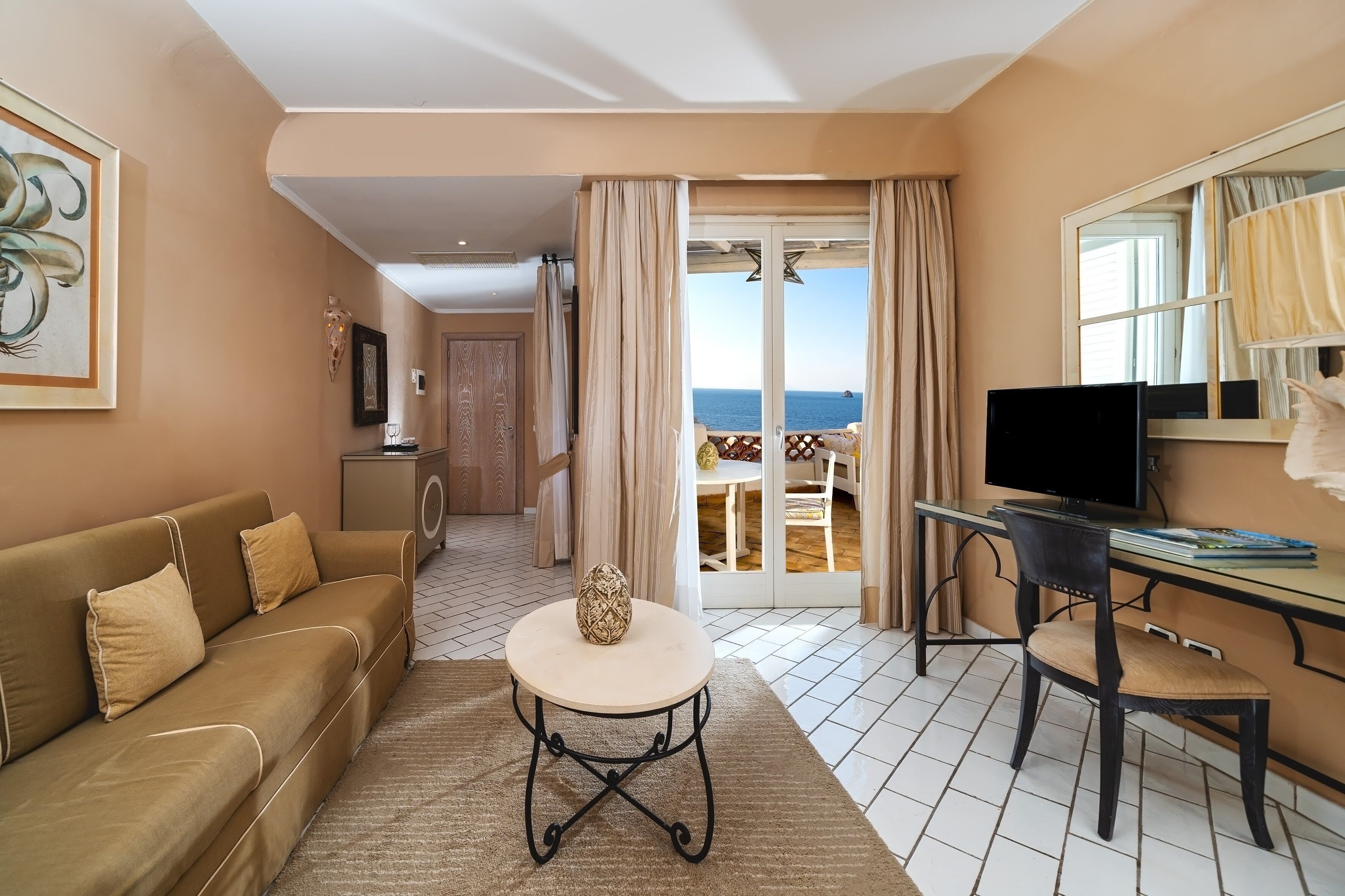 A living room with a sofa, a round table, a TV, and a view of the sea through a glass door.