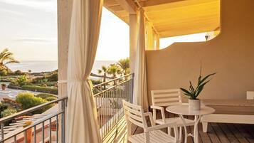 A balcony with white chairs and a table, a potted plant, and a view of the sea and gardens.