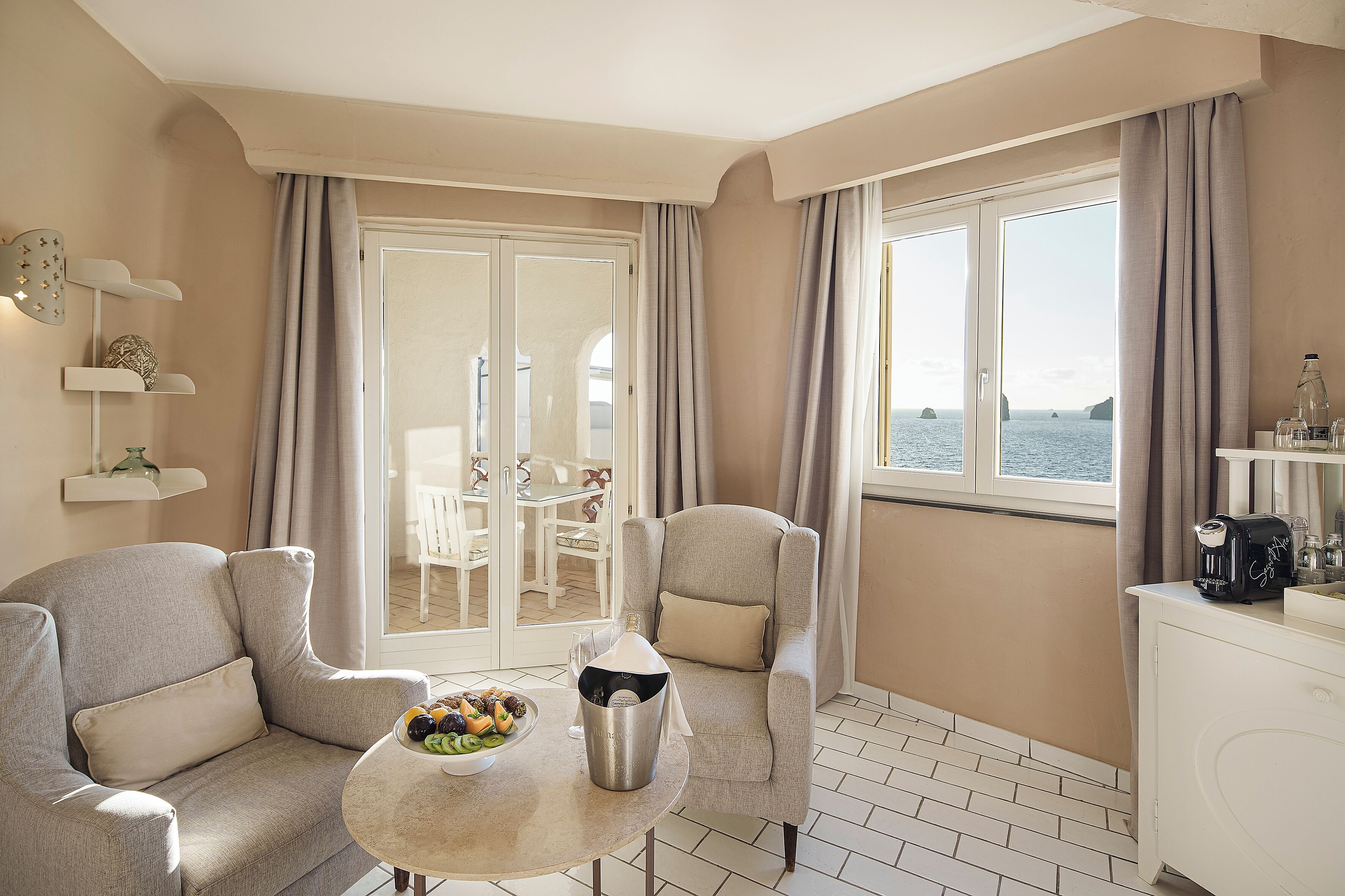 A room with a sofa, a table with a fruit bowl, a coffee maker, and a view of the sea through large windows.
