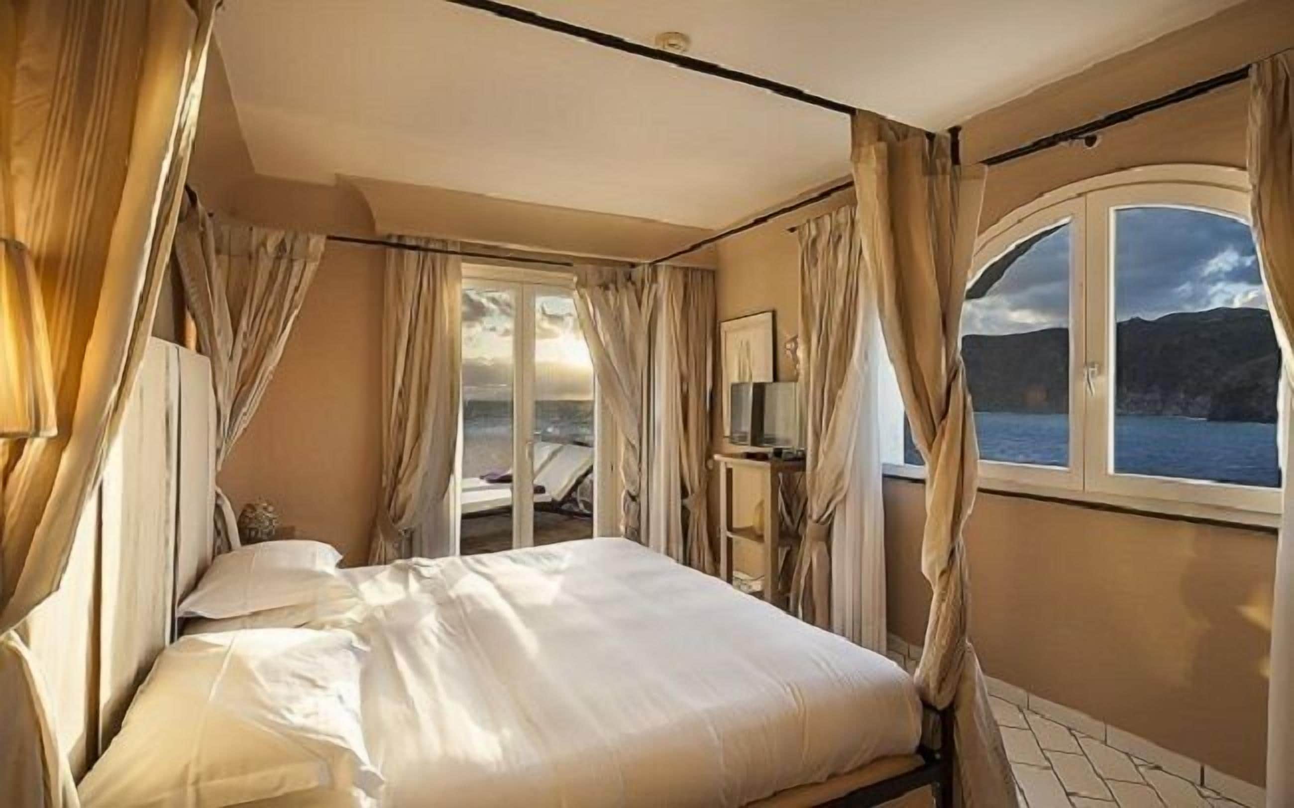 Une chambre à coucher avec un lit, des rideaux, une fenêtre donnant sur la mer et un tableau accroché au mur.
