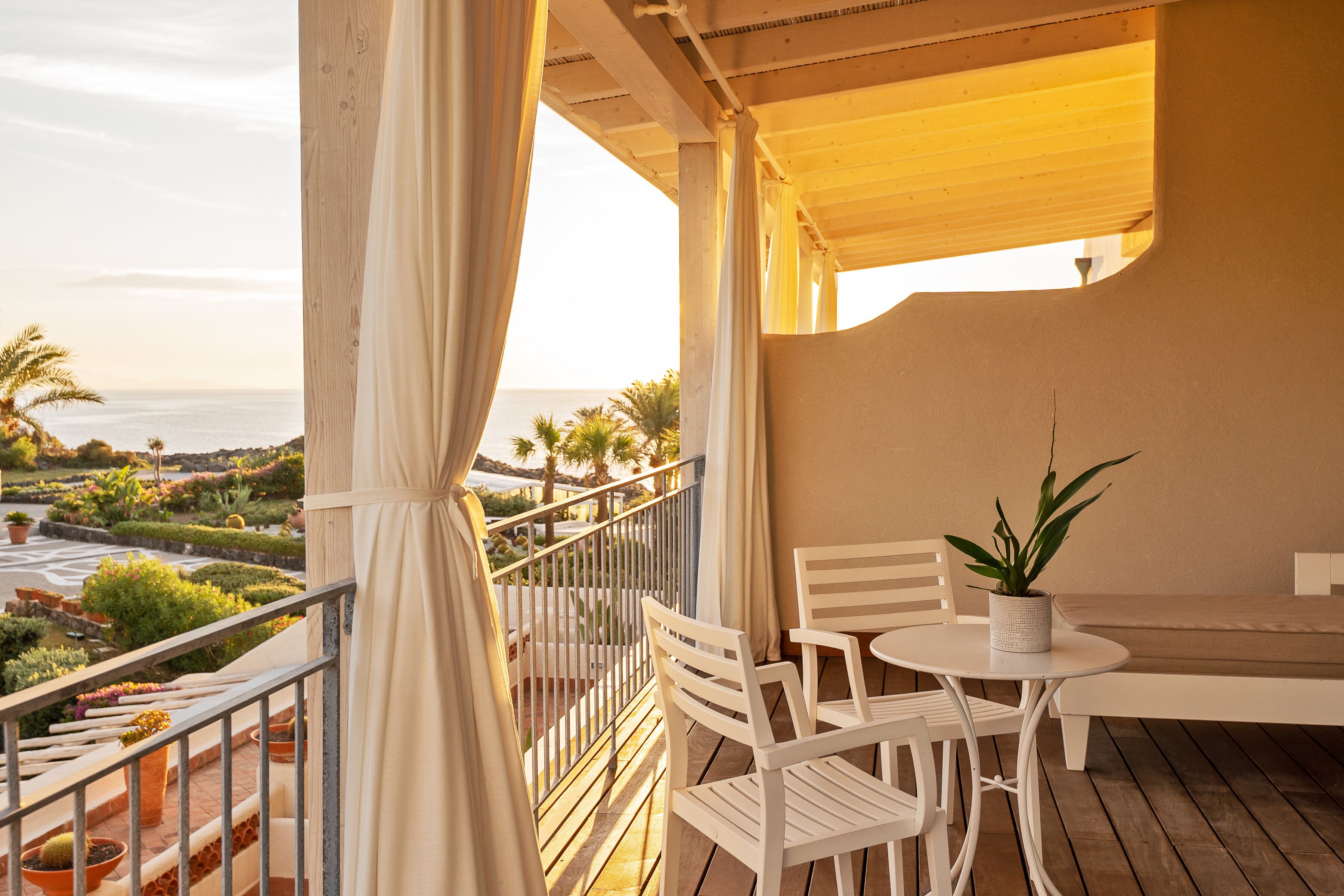 A balcony with white chairs and a table, overlooking a landscaped garden and the sea.