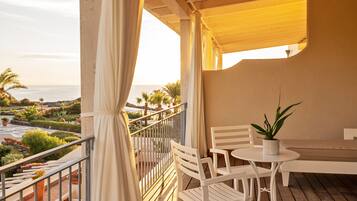 A balcony with white chairs and a table, overlooking a landscaped garden and the sea.