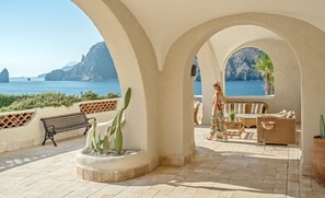 Relax in a serene outdoor space with a clear view of the sea and distant mountains, featuring a sheltered area with a bench, sofa, and potted cactus.