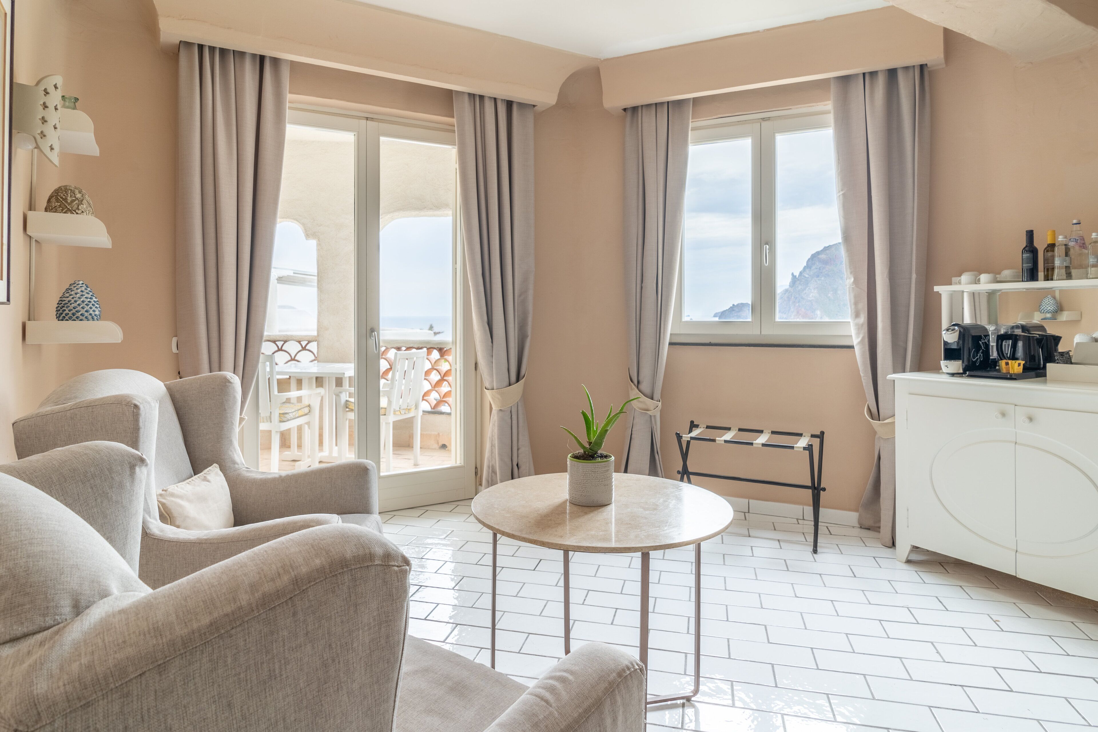 A modern living room with a round table, two armchairs, a potted plant, and a view of the sea through large windows.