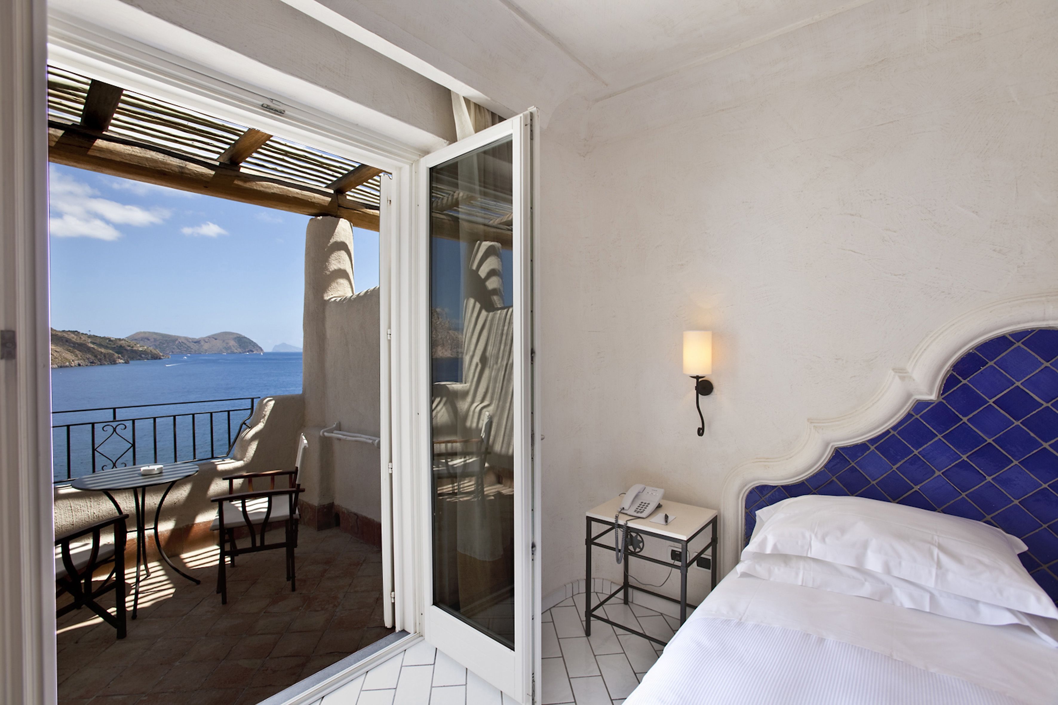 Quarto com cama, mesa de cabeceira, cadeira e mesa. Possui varanda com vista para o mar.
