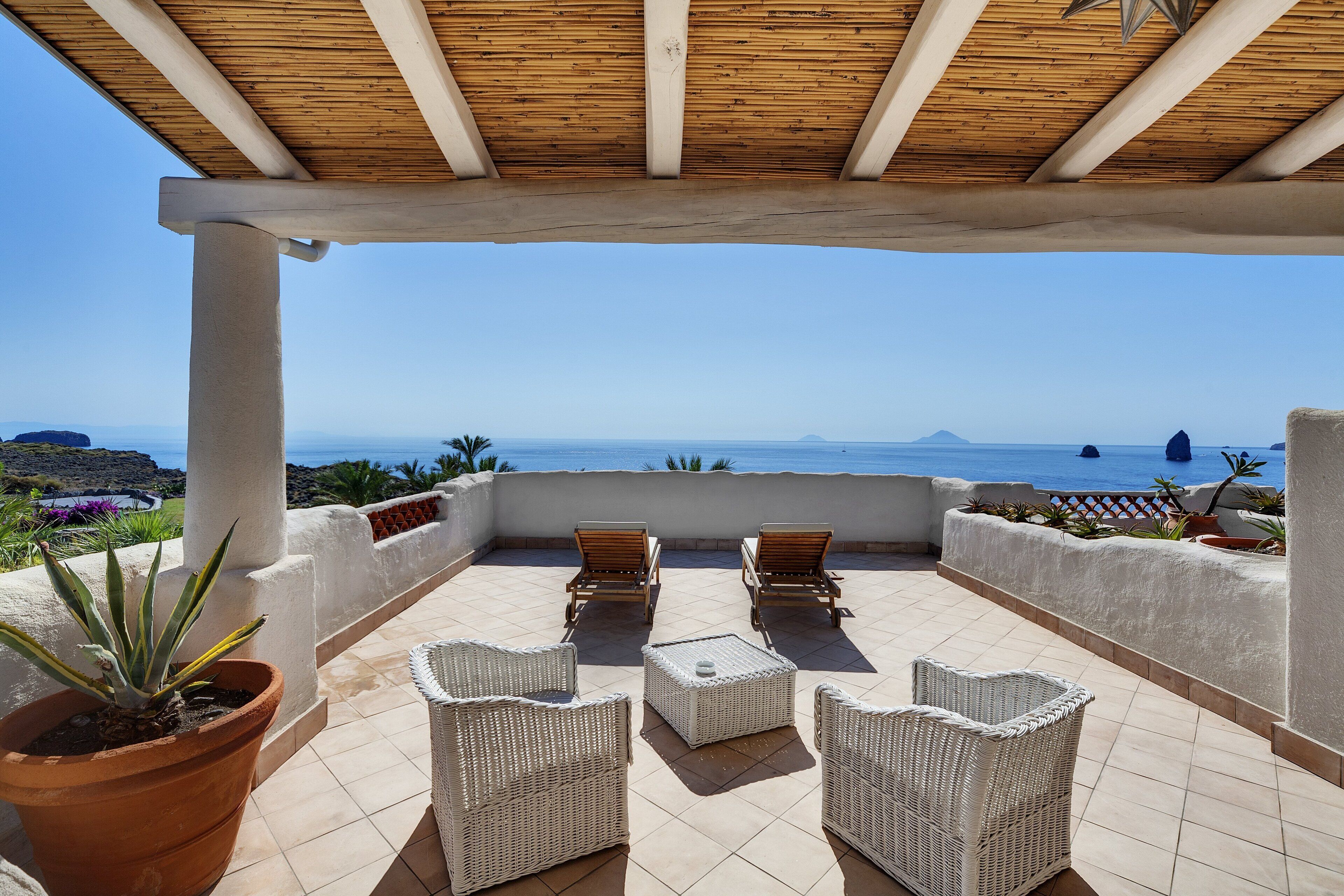 A terrace with wicker furniture and potted plants, overlooking a coastal view.