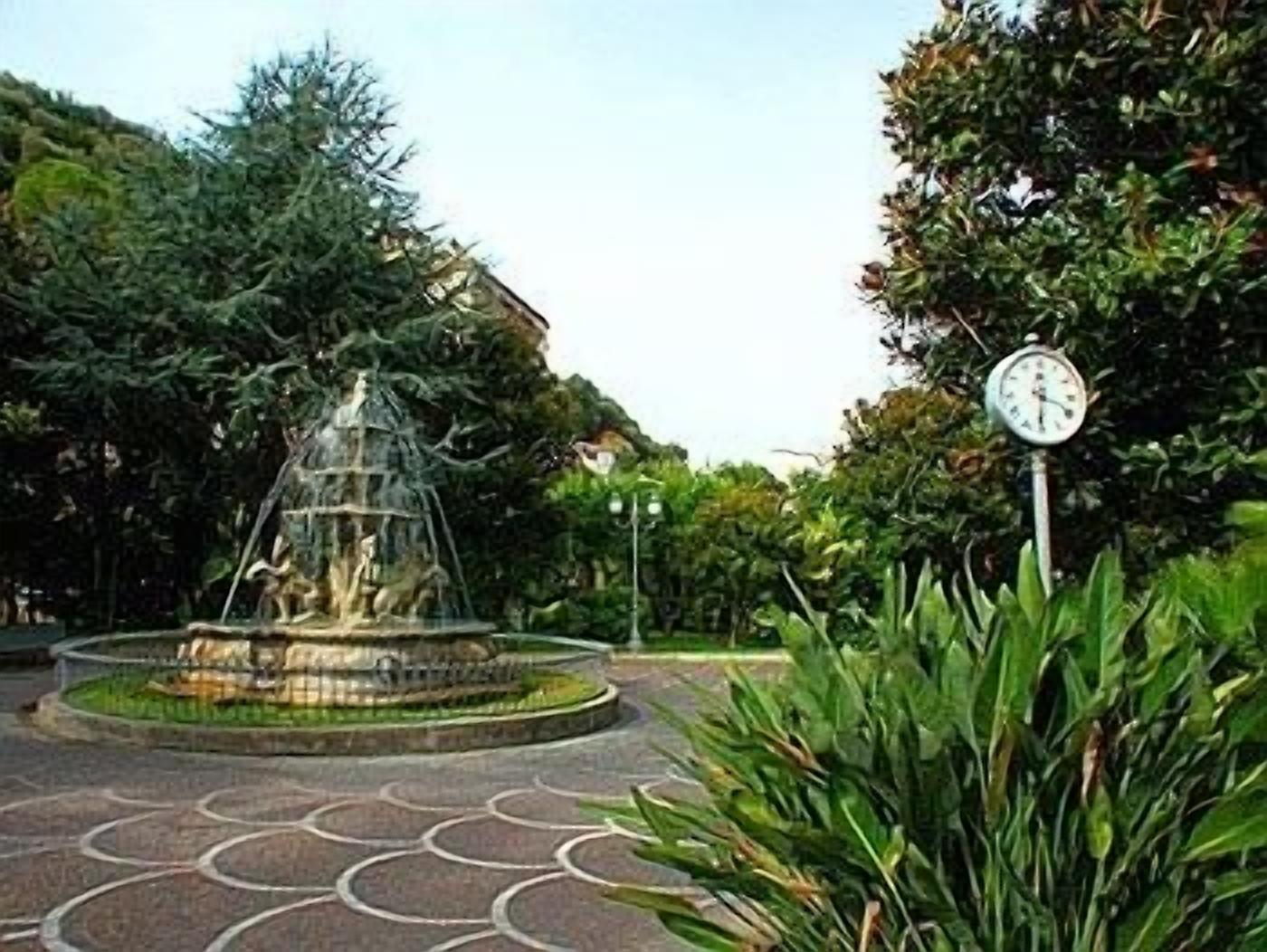 Een ronde fontein met een klok, omringd door groen en een geplaveid gebied.