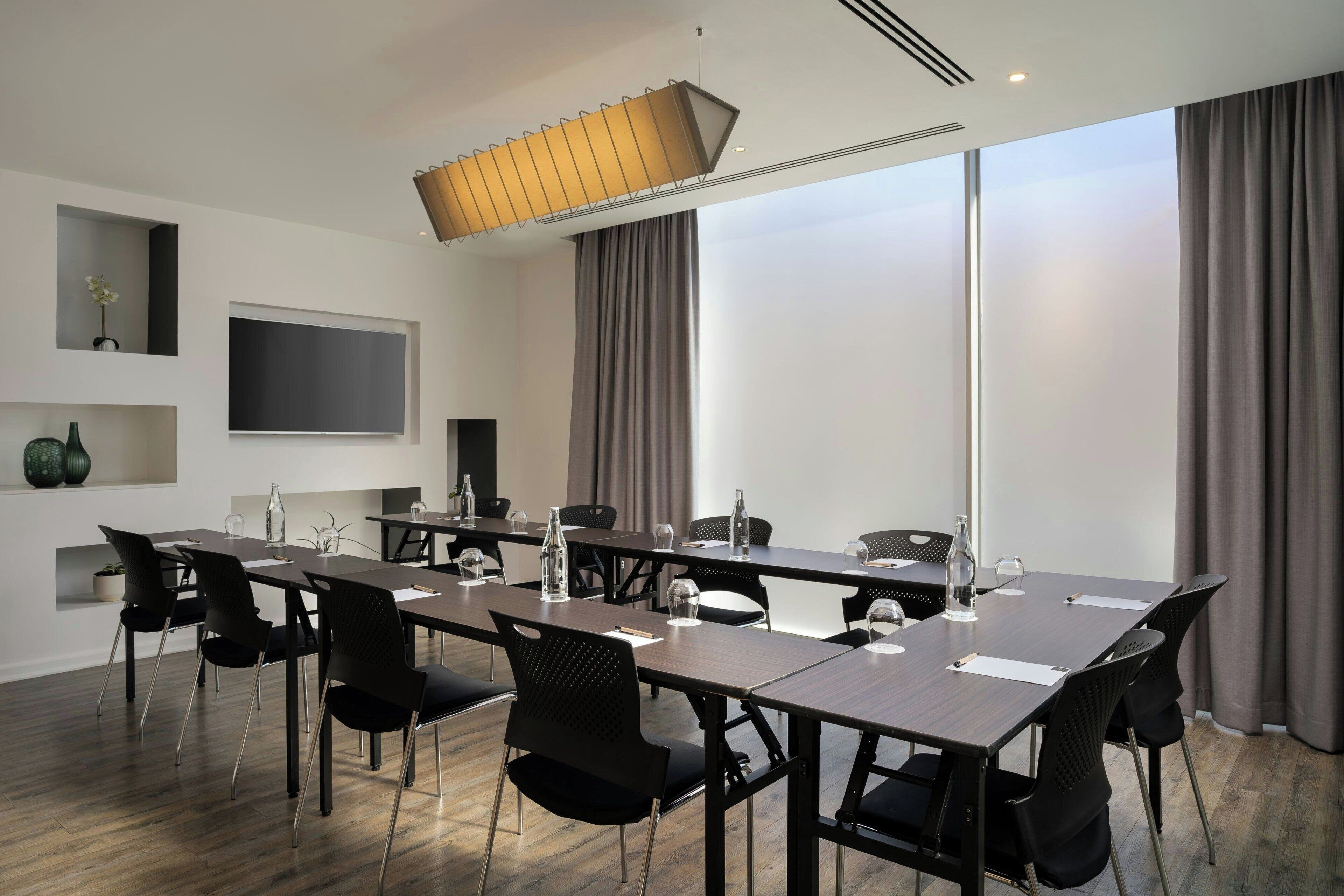 Een vergaderruimte met een lange tafel, zwarte stoelen, een flatscreen-tv en een groot raam met gordijnen.