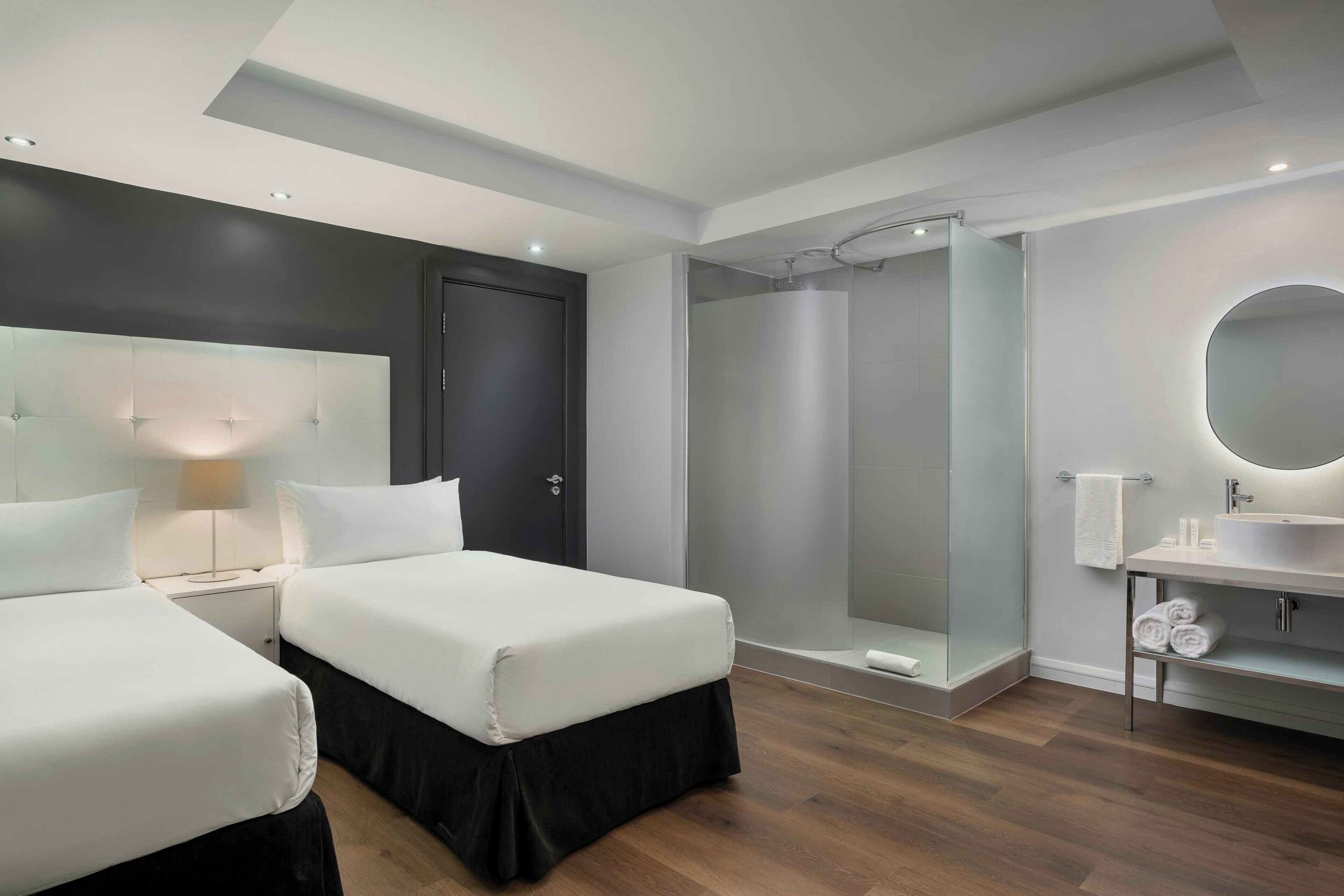 A modern hotel room with two beds, a shower, a vanity with a mirror, and a bedside table with a lamp.