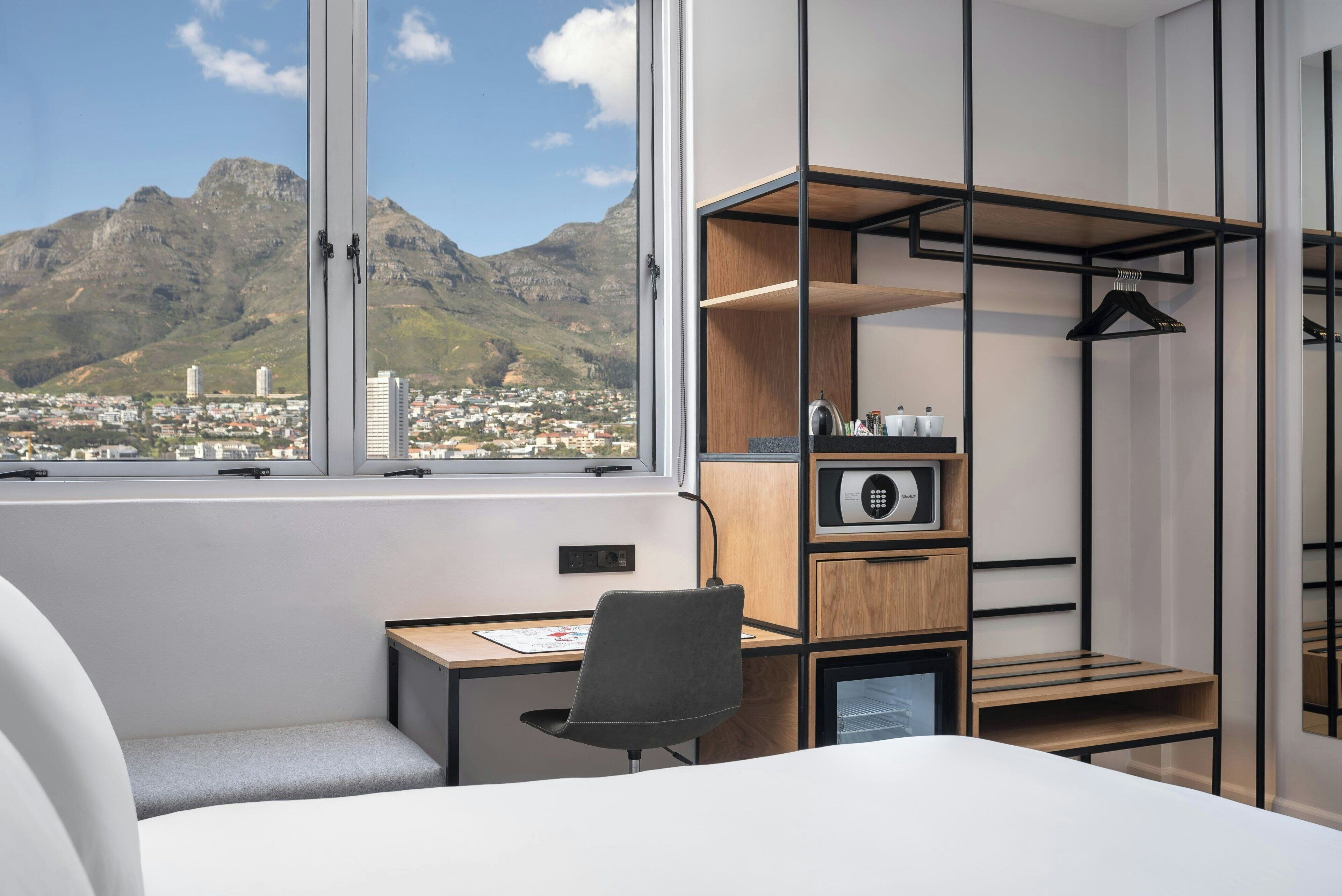 A hotel room with a bed, a desk, and a chair. There is a large window with a view of mountains and a city.