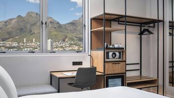A hotel room with a bed, a desk, and a chair. There is a large window with a view of mountains and a city.