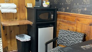 Una kitchenette pequeña equipada con microondas, cafetera y refrigerador. Hay un bote de basura y una silla con un cojín estampado.
