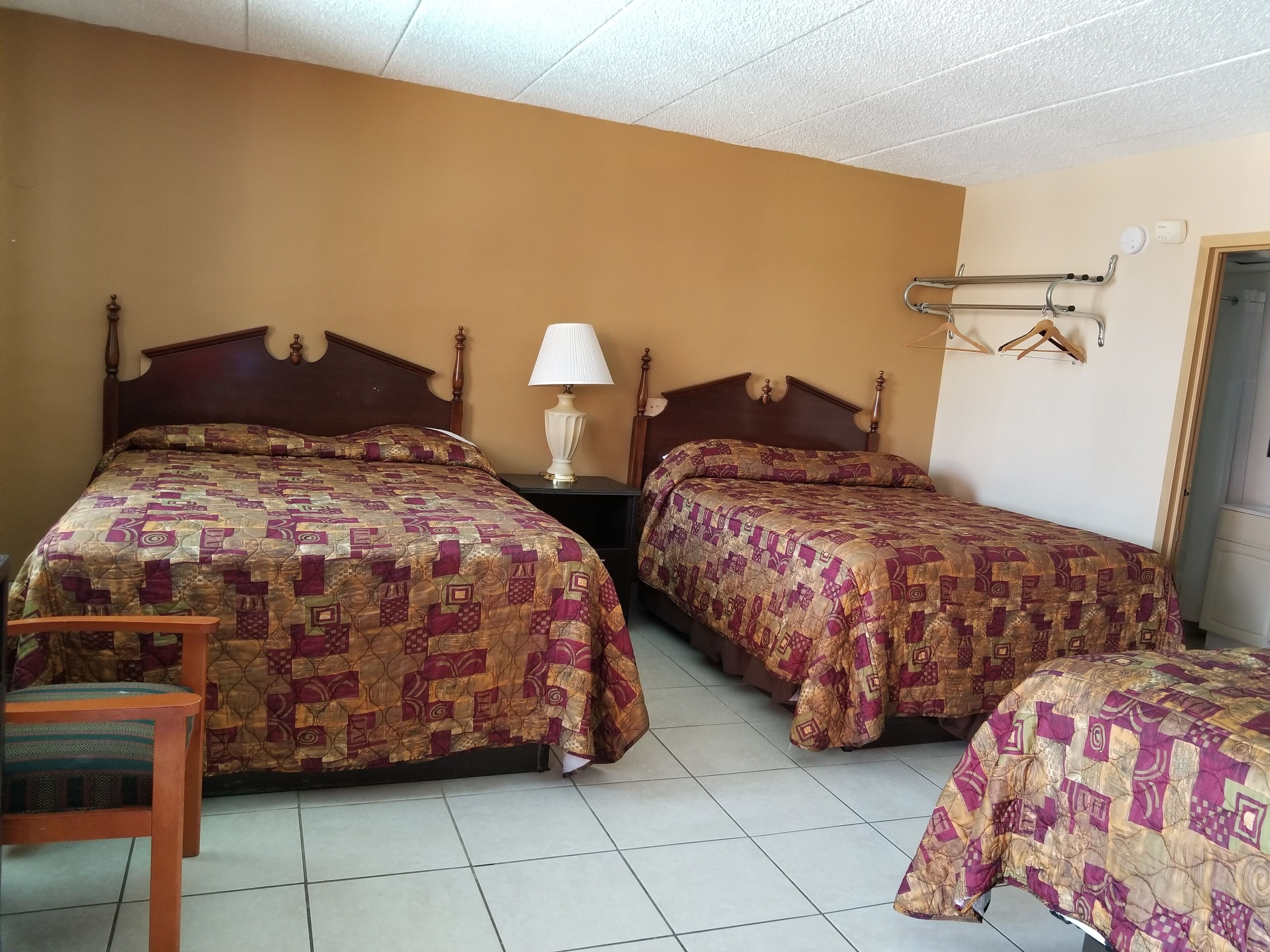 A hotel room with two beds, a nightstand with a lamp, a chair, and a coat rack.
