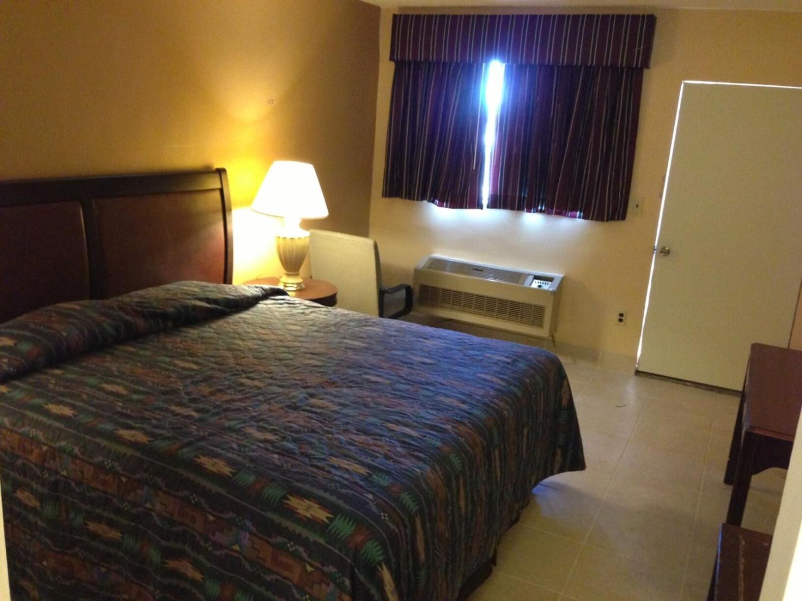 A hotel room with a bed, a nightstand, a lamp, an air conditioning unit, and a door.