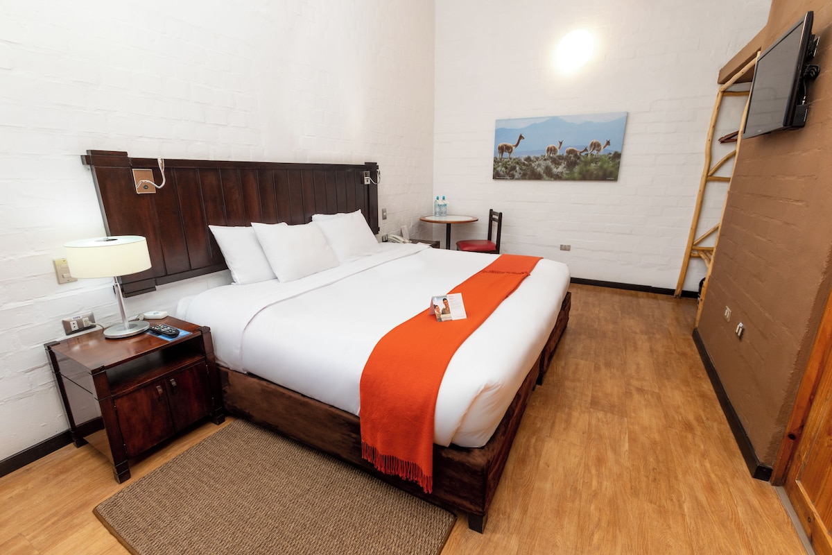 Hotelkamer met een houten bed, witte beddengoed, een klein nachtkastje met een lamp, een rode plaid en een aan de muur bevestigde televisie. Er hangt ook een schilderij van een landschap.